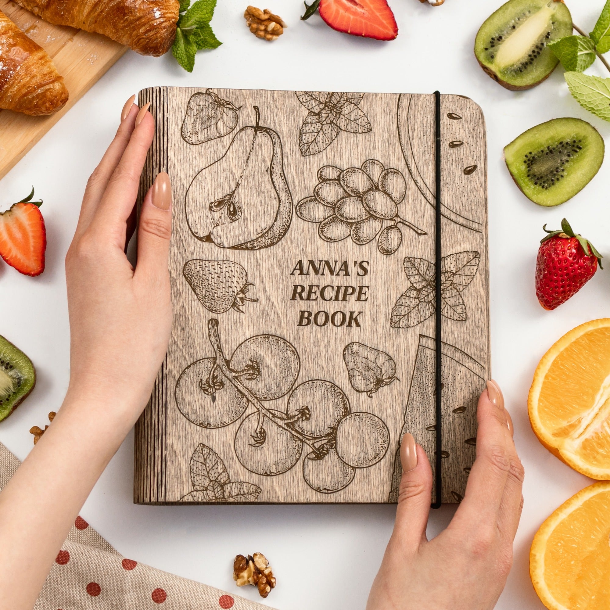 Wooden recipe book titled 'Anna's Recipe Book' surrounded by fruits and a hand holding it.