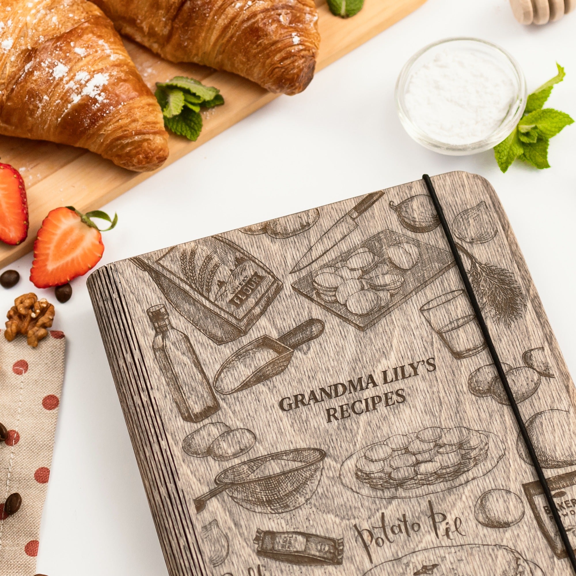 Notebook titled 'Grandma Lily's Recipes' with kitchen-themed design on a table with croissants and strawberries.