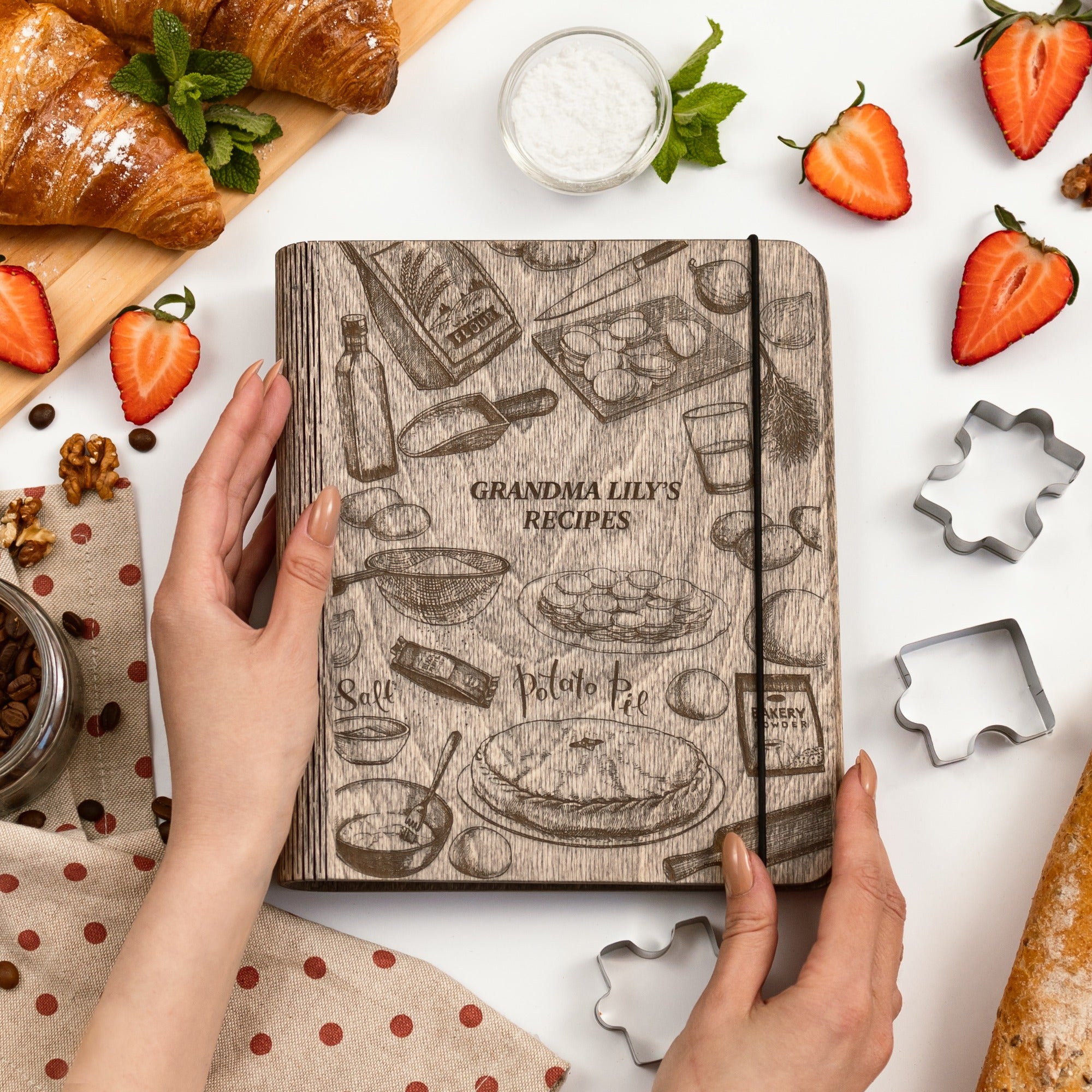 Hand holding a recipe book titled 'Grandma Lily's Recipes' with kitchen items and food around it.