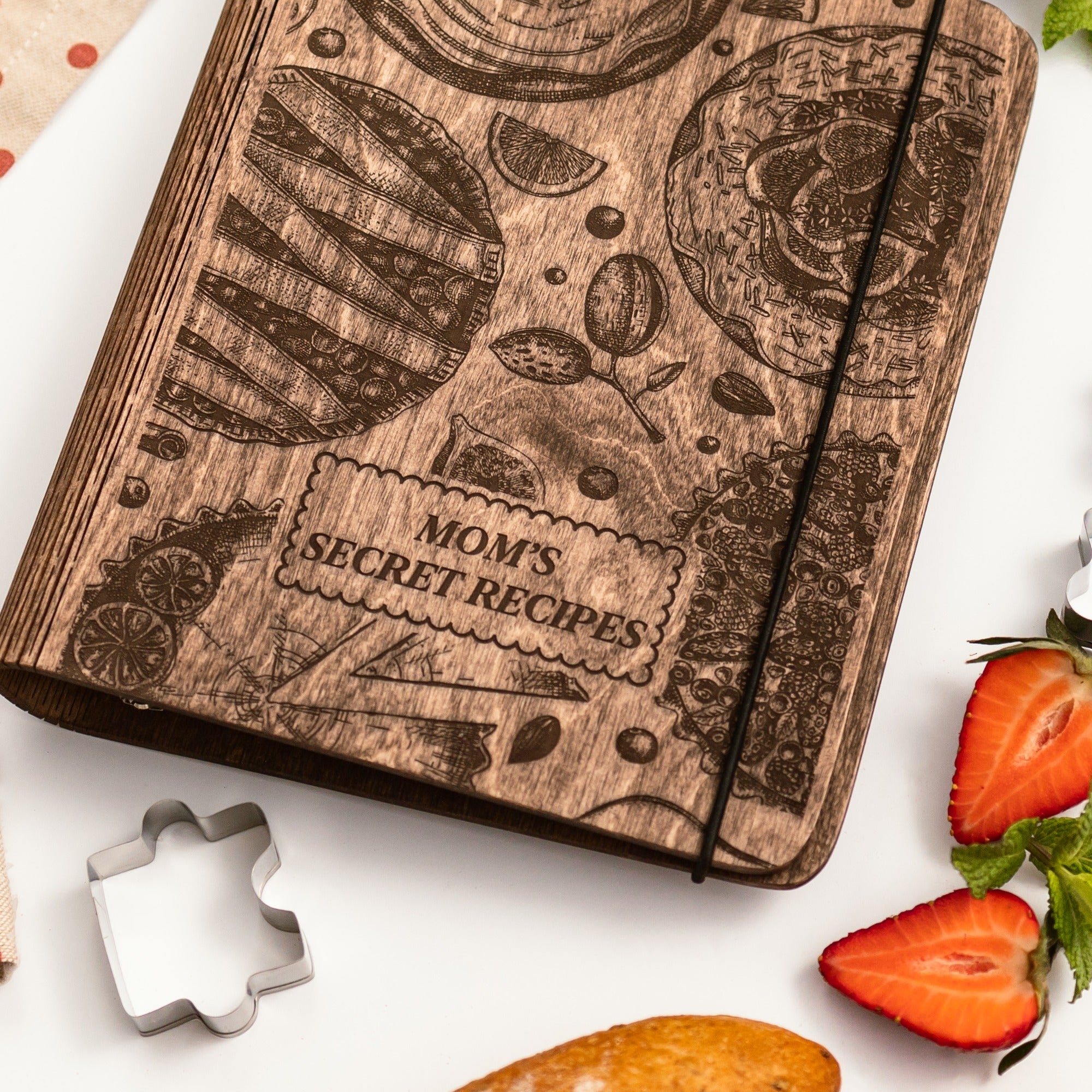 Wooden recipe book with engraved text and illustrations on a white surface with strawberries and a cookie cutter.