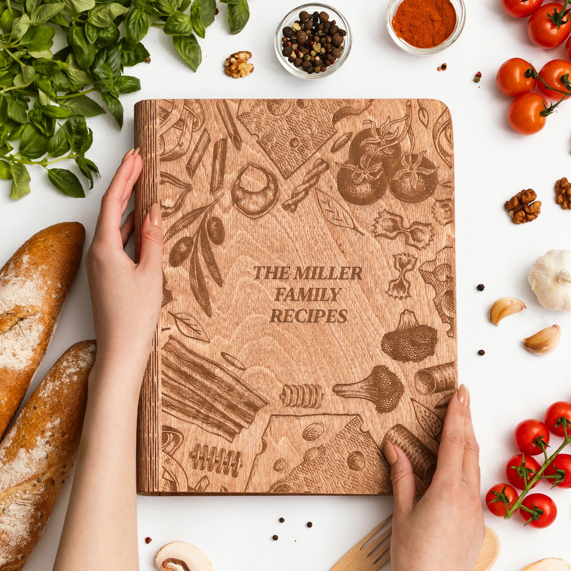 Wooden recipe book titled 'The Miller Family Recipes' held by hands with ingredients like bread, tomatoes, and herbs on a white background.