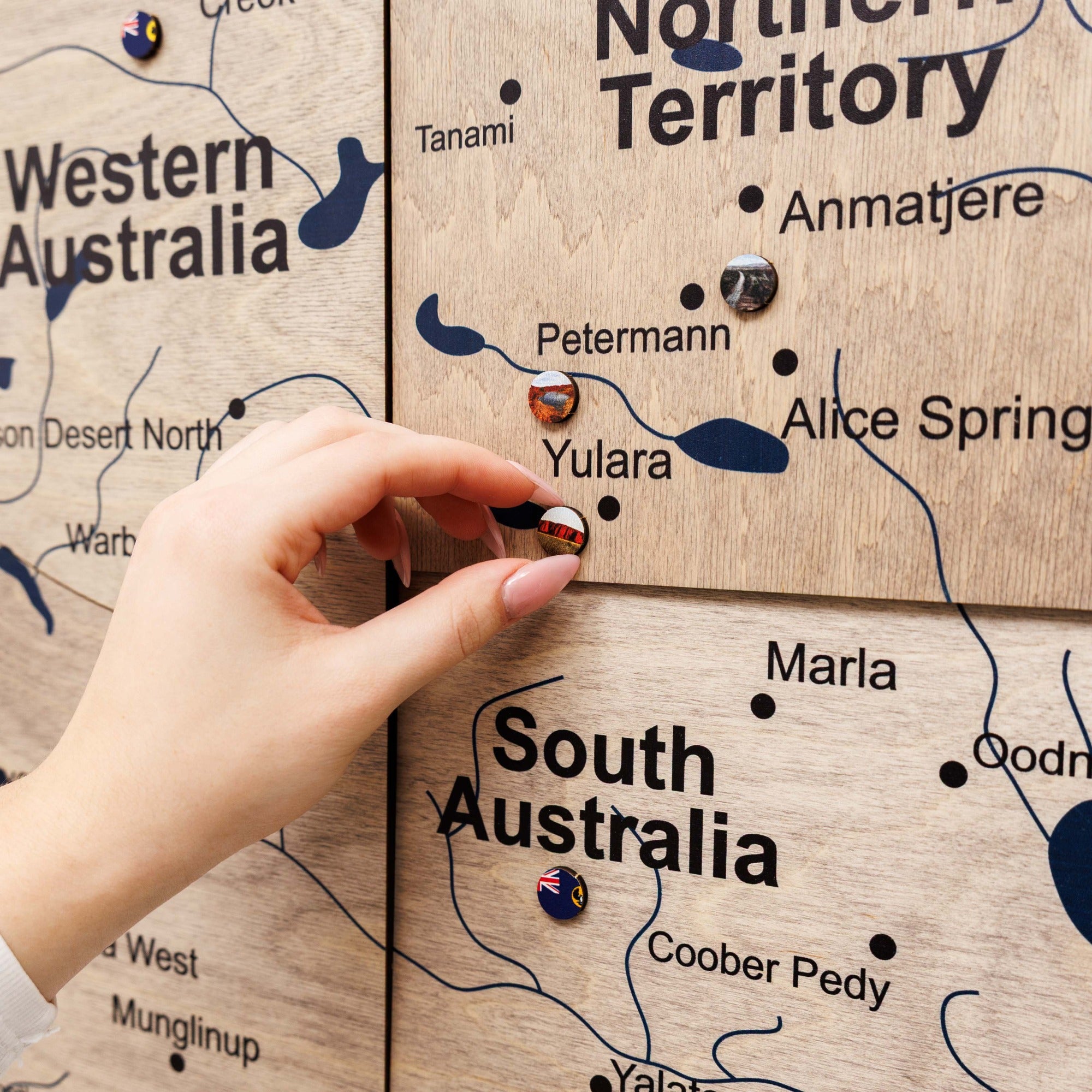 Hand placing a pin on a wooden map of Australia, highlighting regions like Western Australia, Northern Territory, and South Australia.