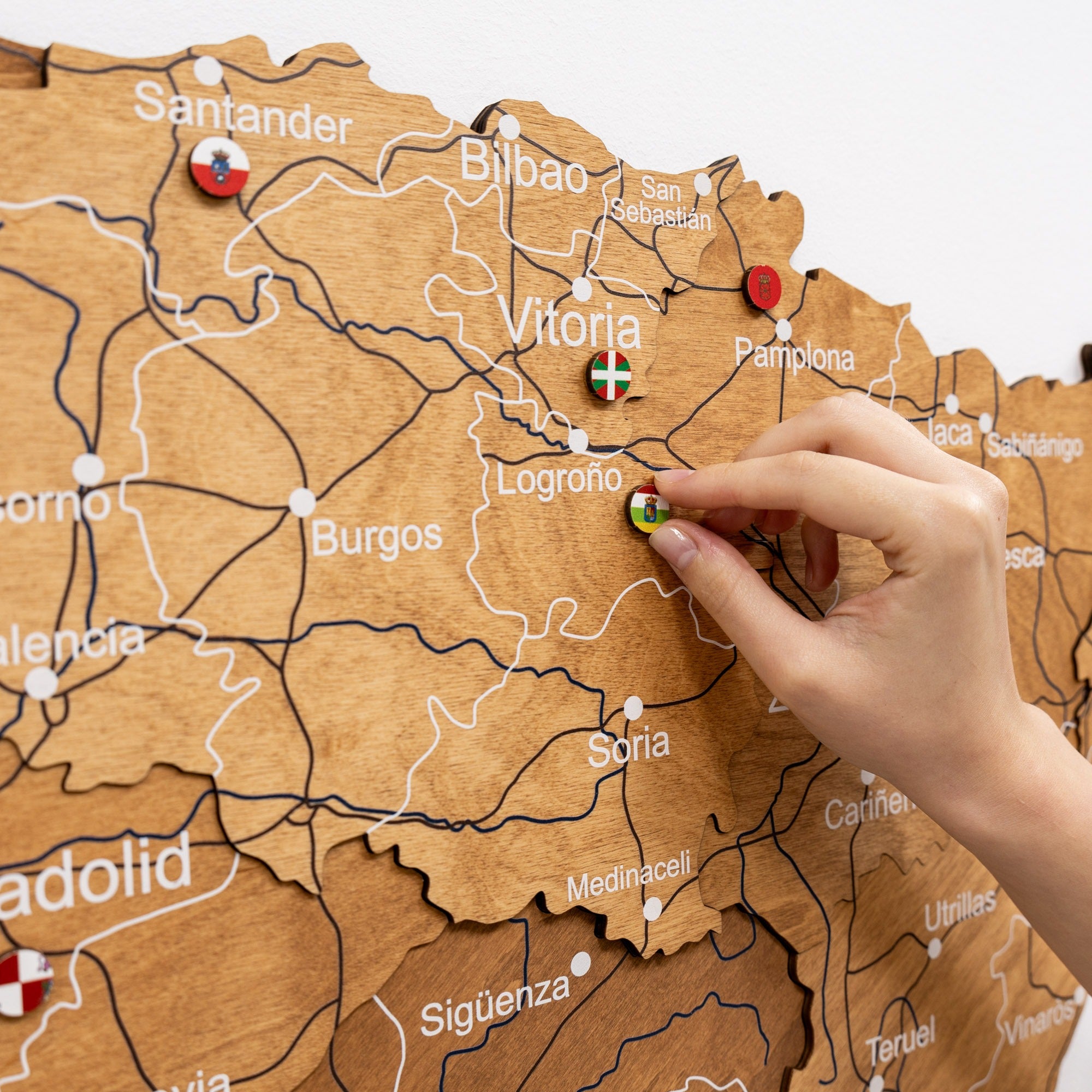 Hand placing a pin on a wooden map of Spain with various cities labeled.