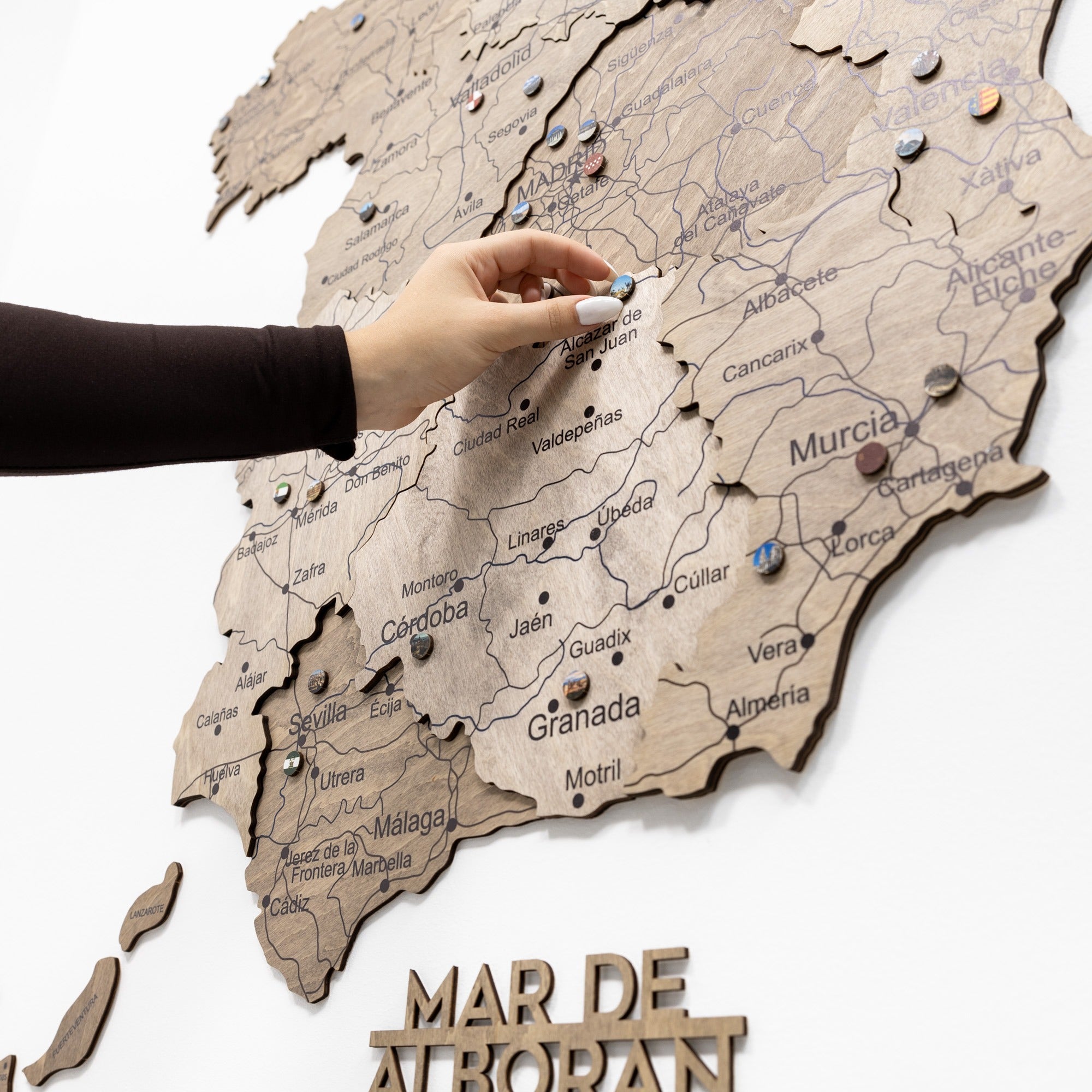 Hand placing a pin on a wooden map of Spain with 'Mar de Atropan' at the bottom.