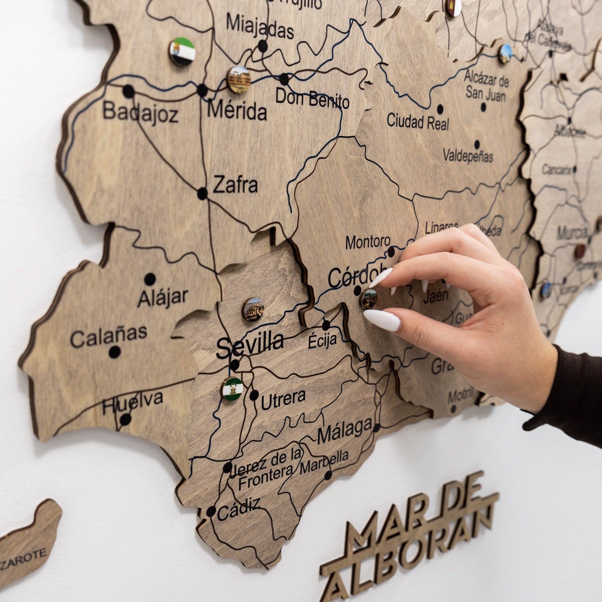 Wooden map of Spain with pins marking locations, hand pointing to Seville.