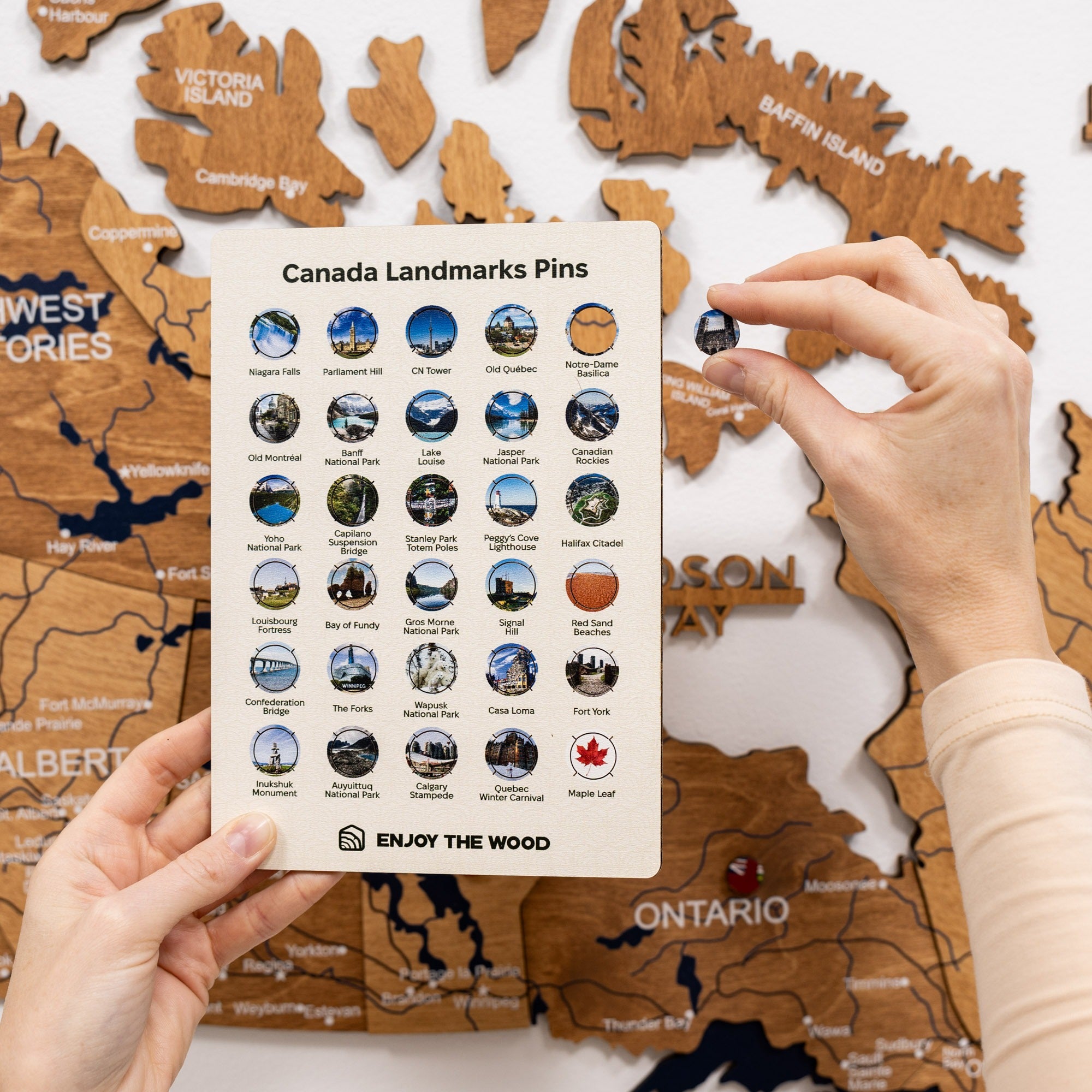 Wooden map of Canada with pins and a card labeled 'Canada Landmarks Pins' held by a person.