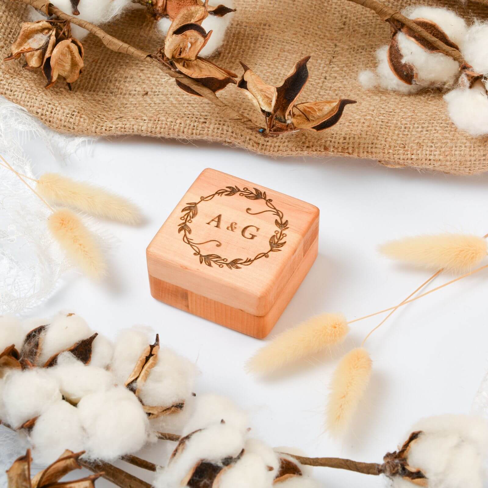 A wooden ring box with engraved initials 'A & G' on the lid, displayed alongside cotton plants and a burlap fabric.