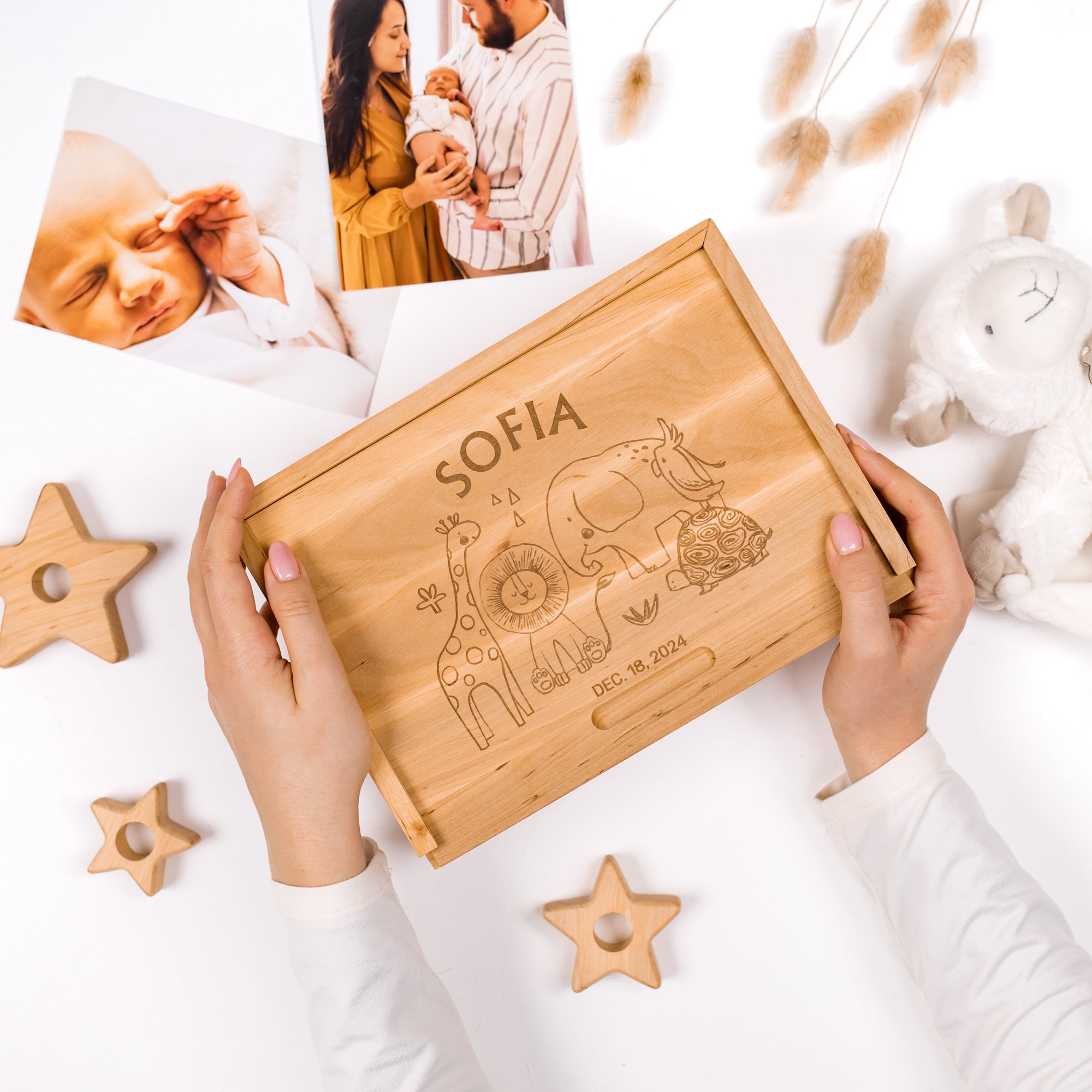 Wooden box with engraved animals held by hands, surrounded by baby items on a light background