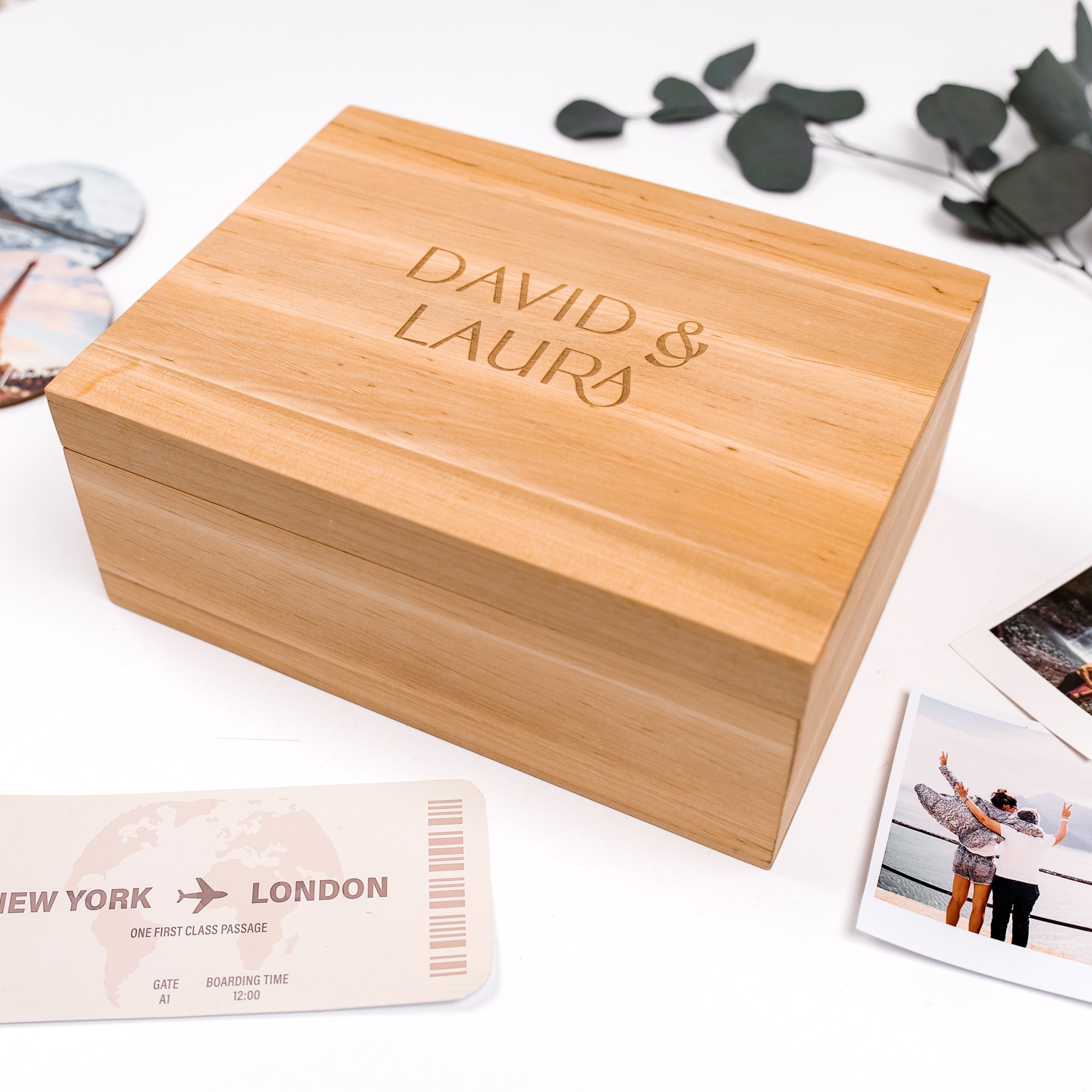 Wooden box with 'DAVID & LAURA' engraving, surrounded by travel-themed items on a white surface.