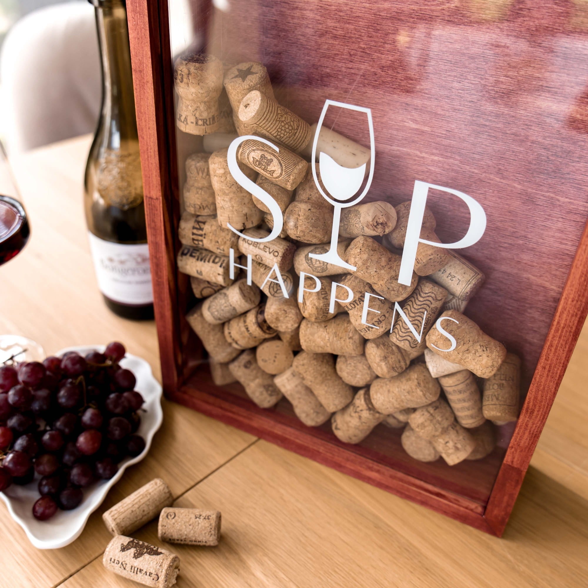 A wooden gift box filled with wine corks, featuring a clear front panel with the white text 'SIP HAPPENS' and a wine glass icon. The box sits on a wooden table beside a bottle of wine, a small dish of dark grapes, and a few loose corks.