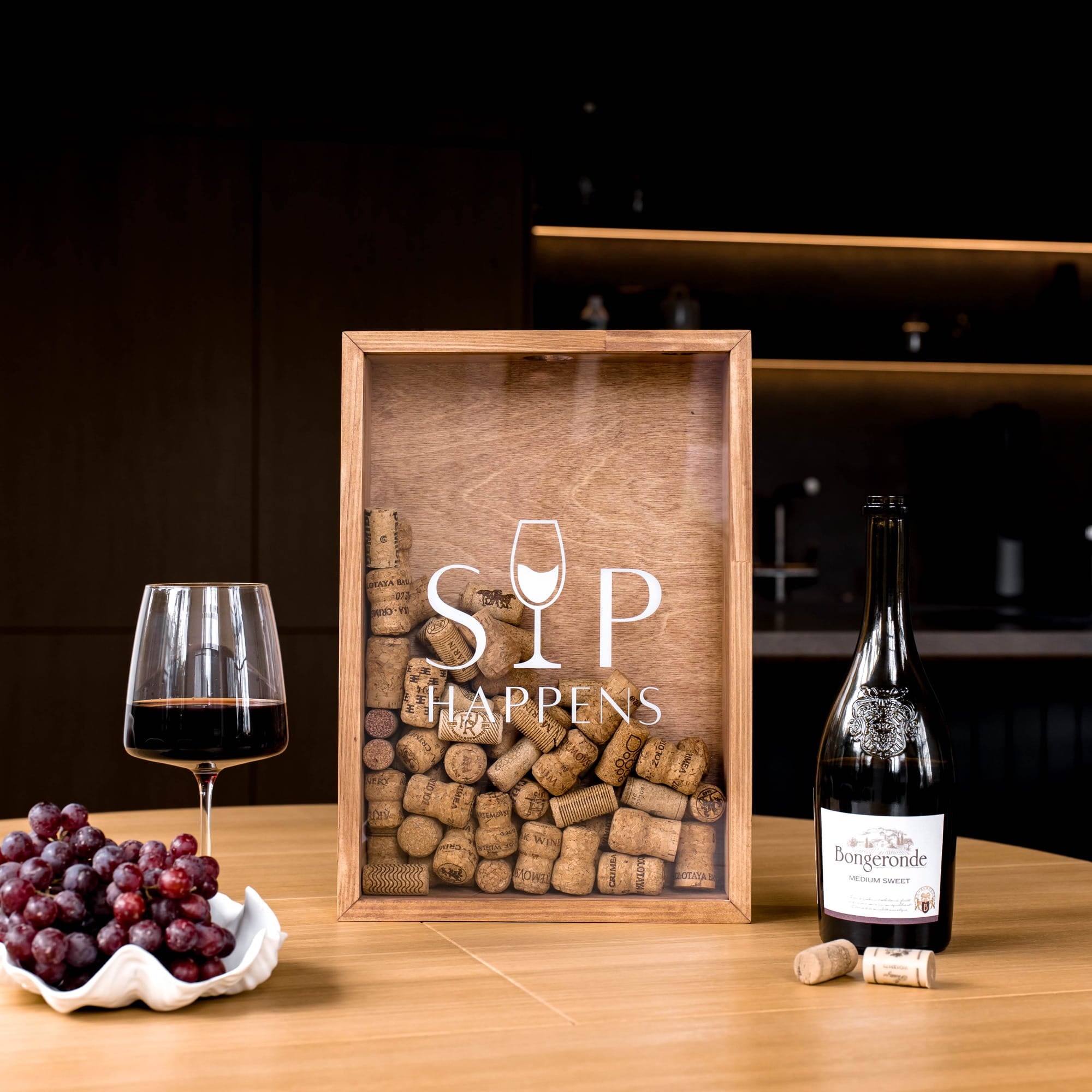 A wooden framed wine-themed decor piece on a wooden table, featuring the words 'SIP HAPPENS' and a wine glass icon over a collage of wine corks.