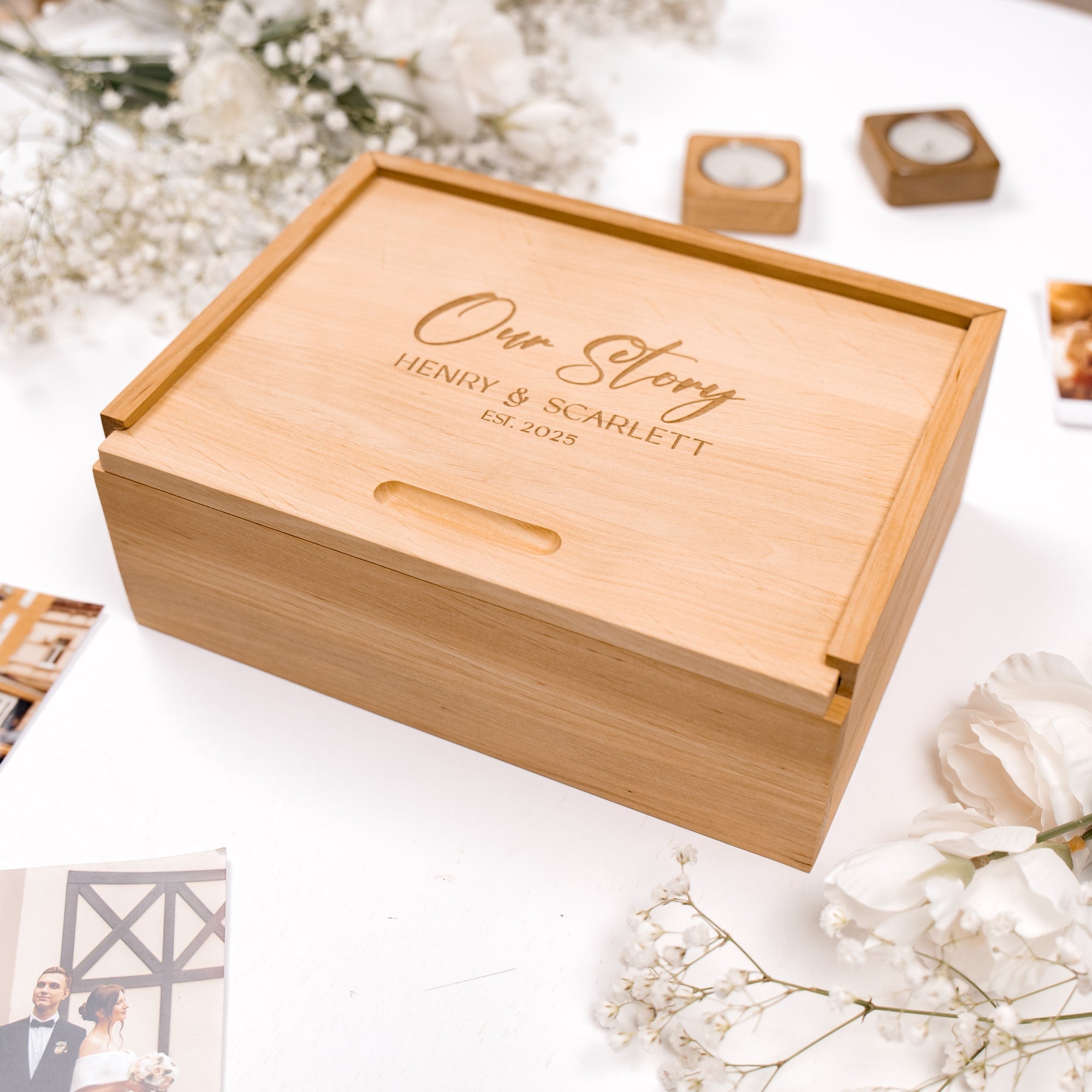Wooden box with 'Our Story Henry & Scarlett' engraving on a white surface with flowers and candles.