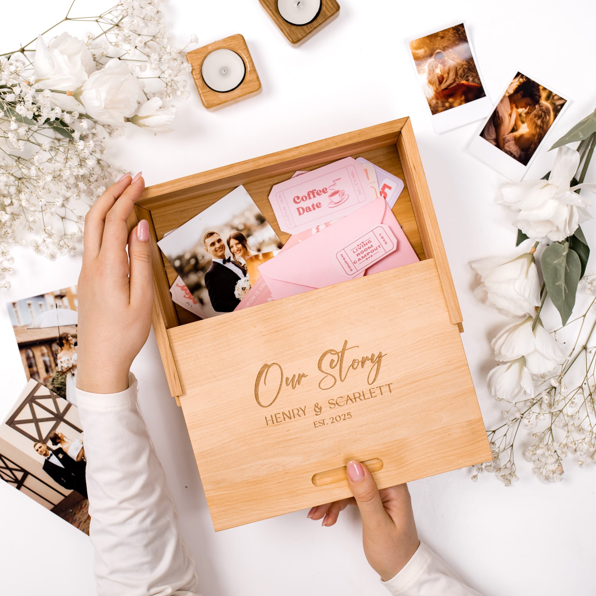 Wooden box with 'Our Story' engraving, held by hands, surrounded by photos and flowers on a white surface.