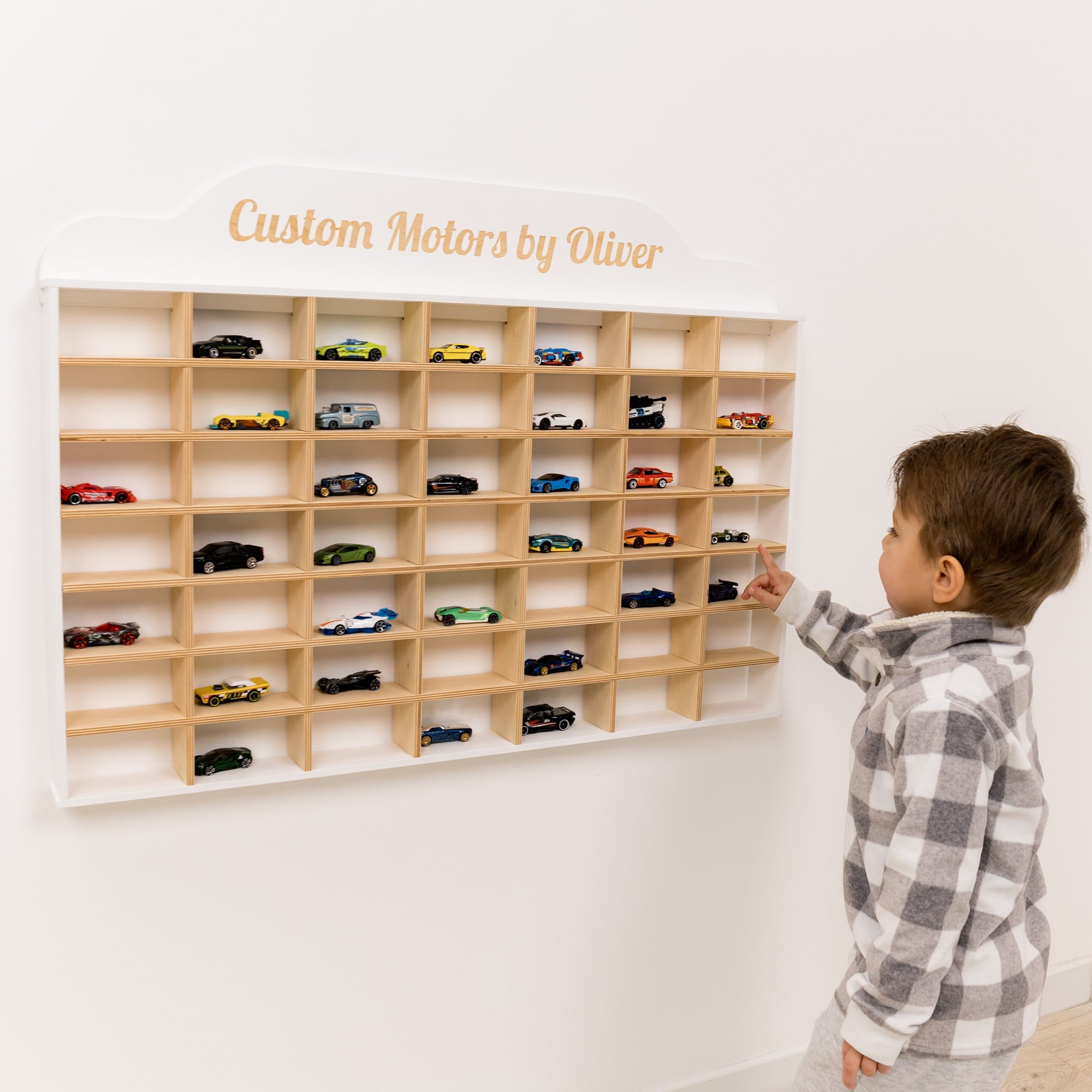 Child interacting with a wooden wall display of toy cars labeled 'Custom Motors by Oliver'.