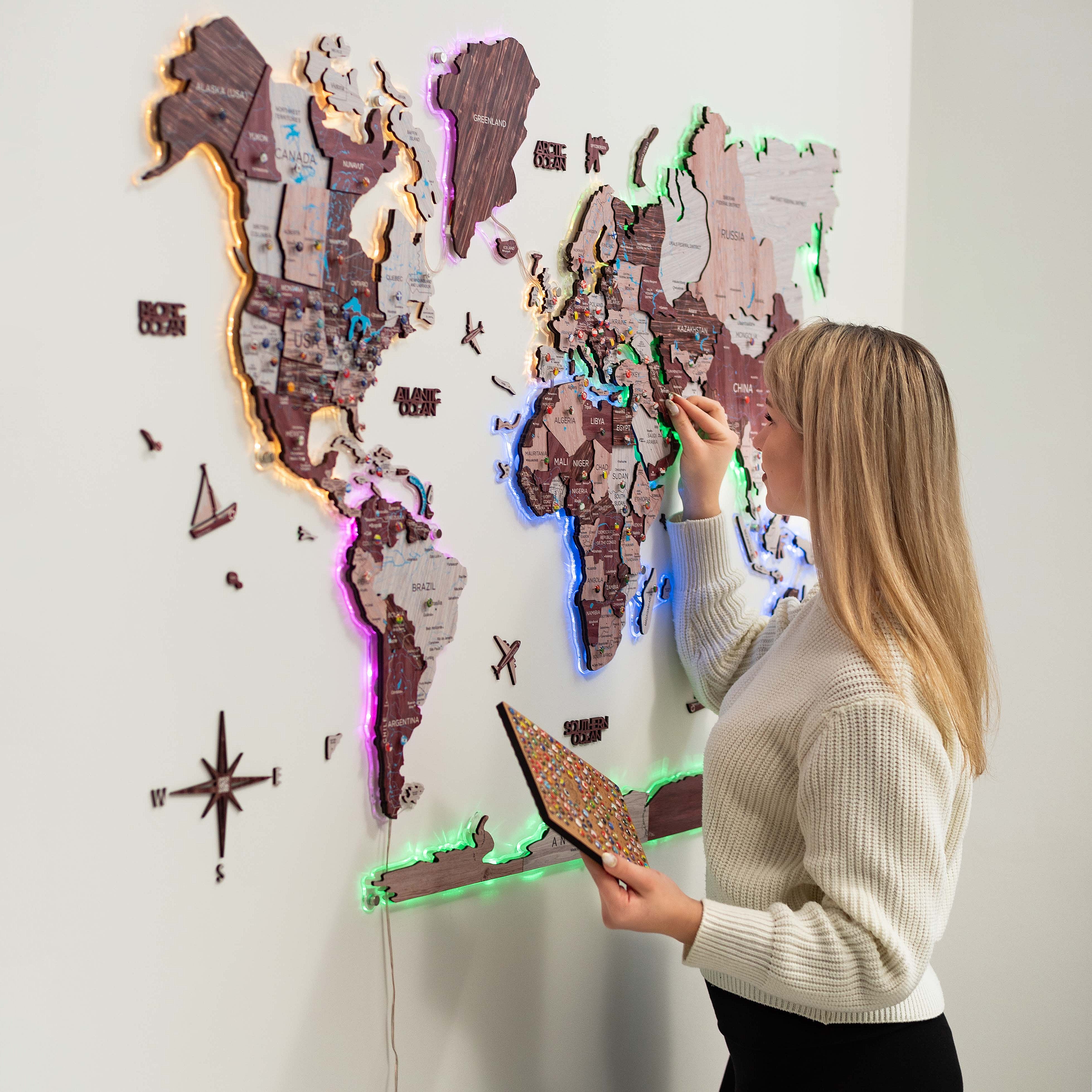 A person is placing a wooden world map with luminous, magnetic features on a wall. The map is illuminated with LED lights and has various colors.