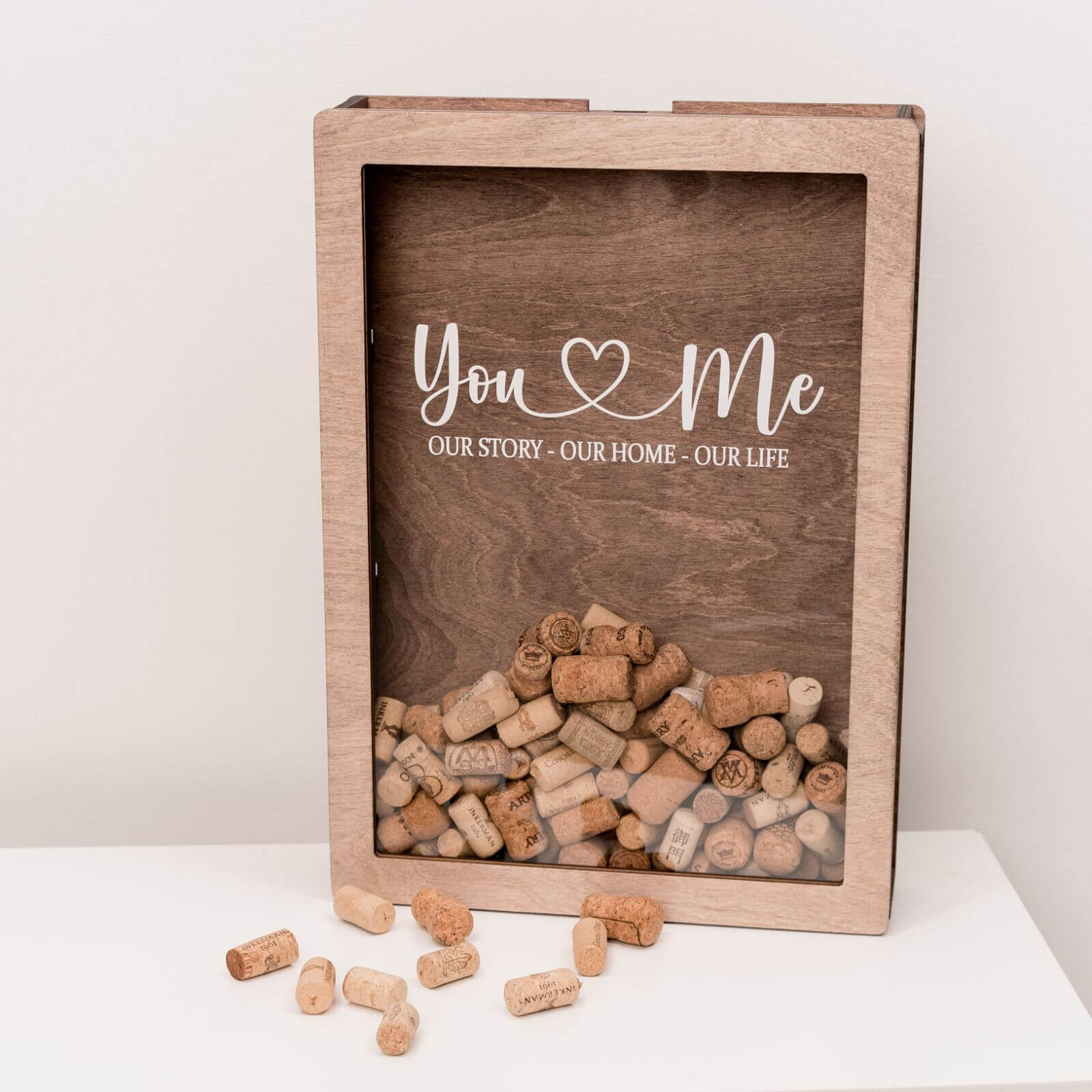 A wooden wine cork box with a glass front displaying text and a wine tap illustration, placed on a table with wine glasses and bottles in the background.
