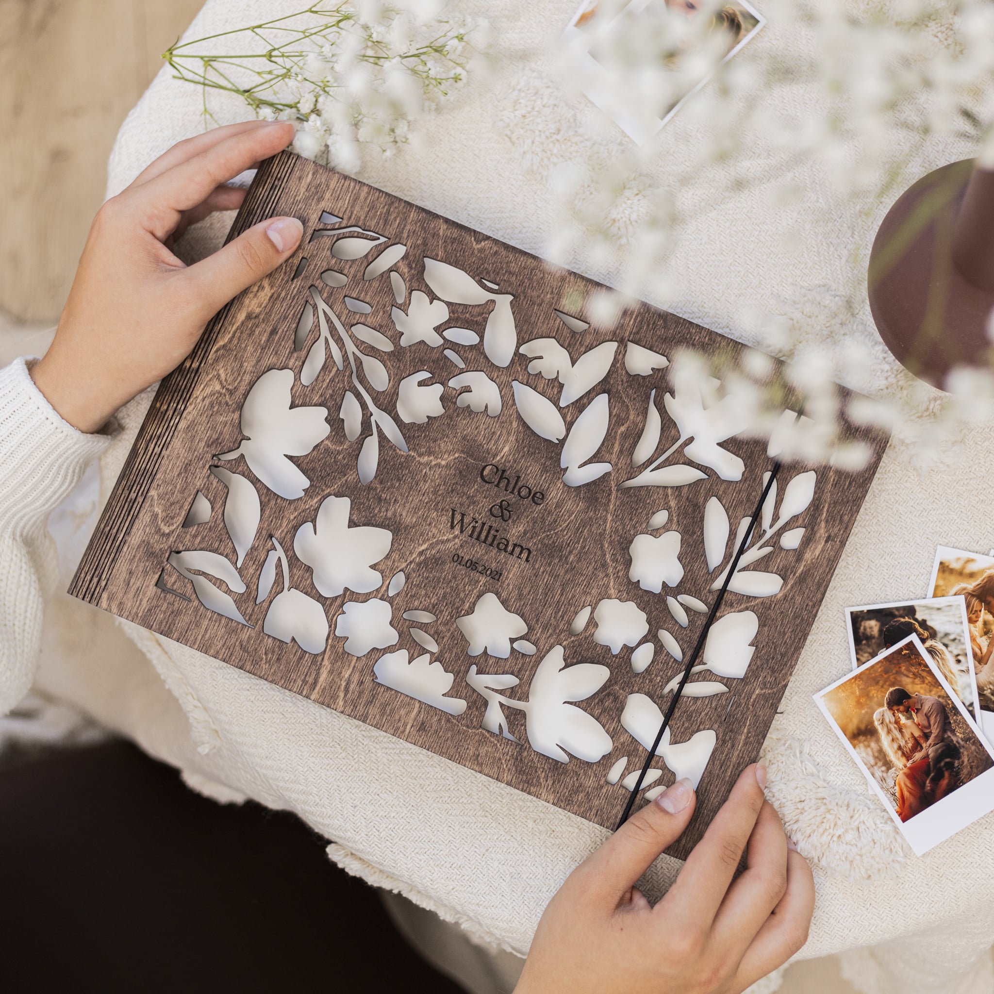 Wooden photo album with floral design and engraved names, held by a person on a soft white surface.