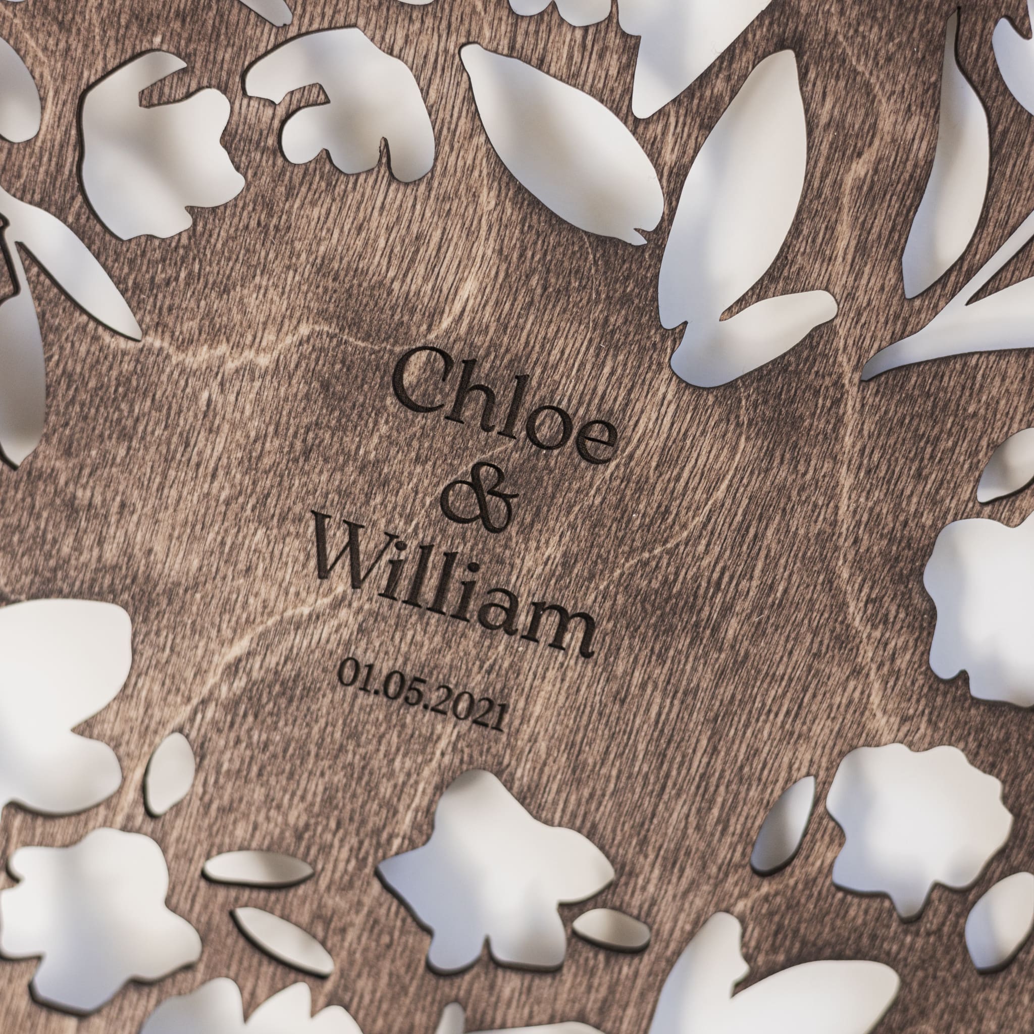 Wooden plaque with engraved names 'Chloe & William' and date '01.05.2021', surrounded by white floral cutouts.