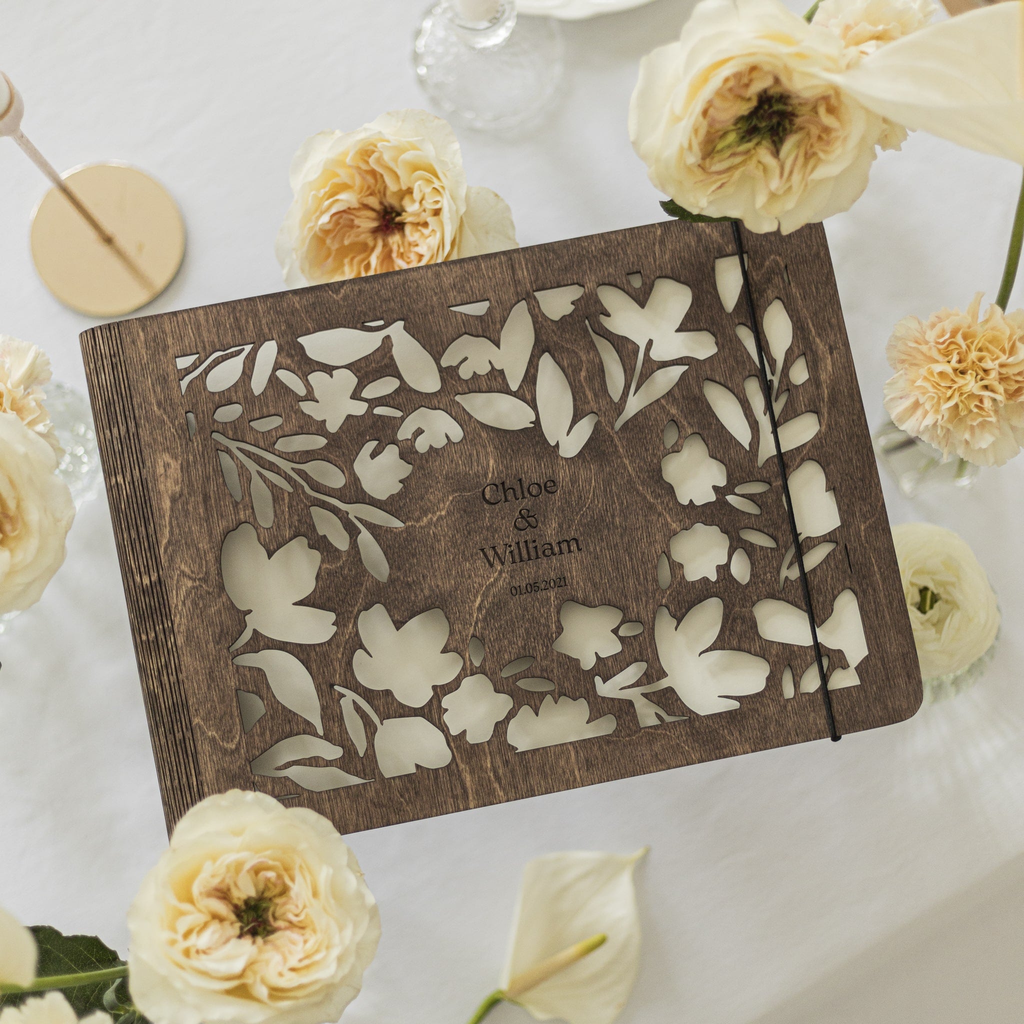 Wooden album with floral cutout design and engraved names, surrounded by white flowers on a light background