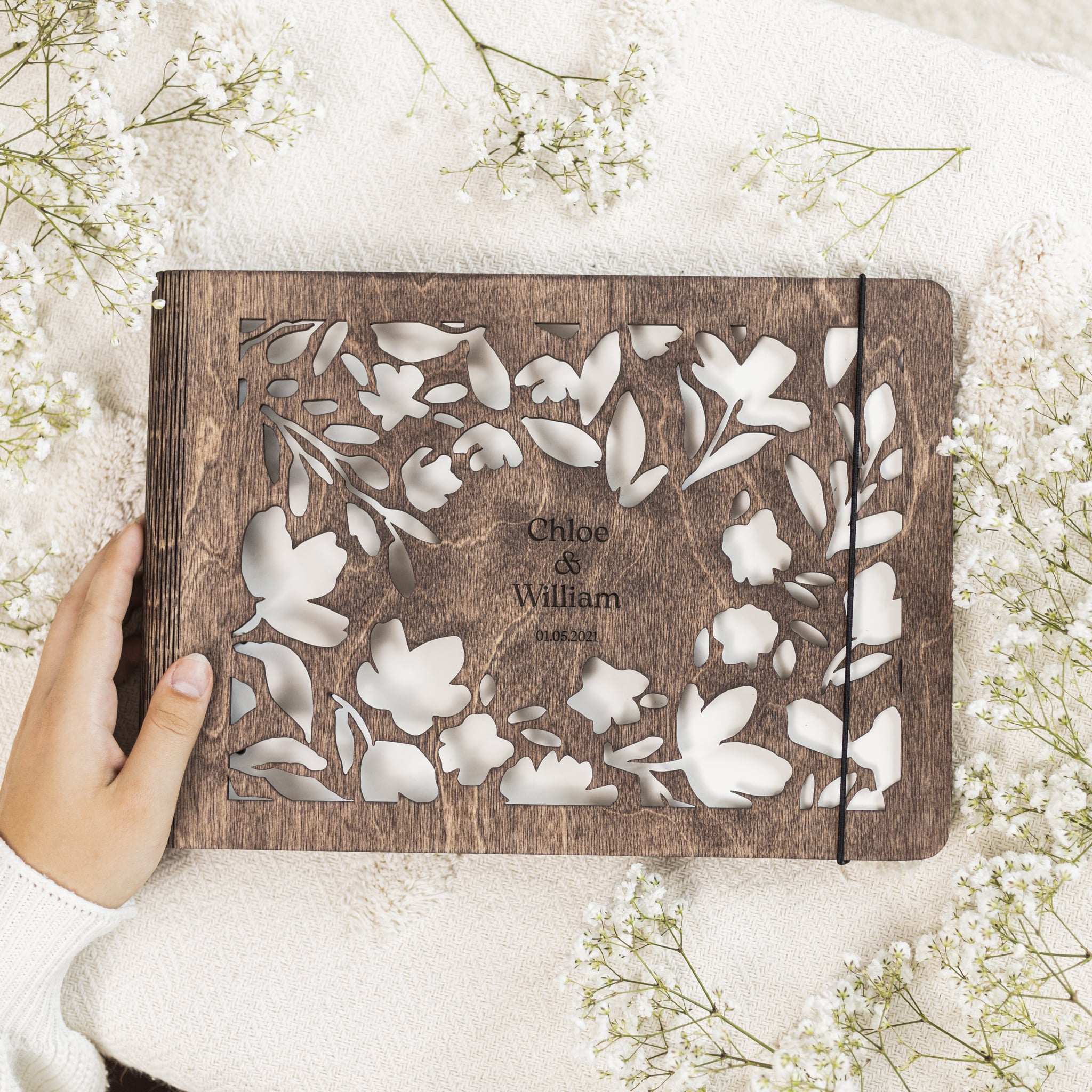 Wooden photo album with floral design held by a hand on a white background with small flowers.