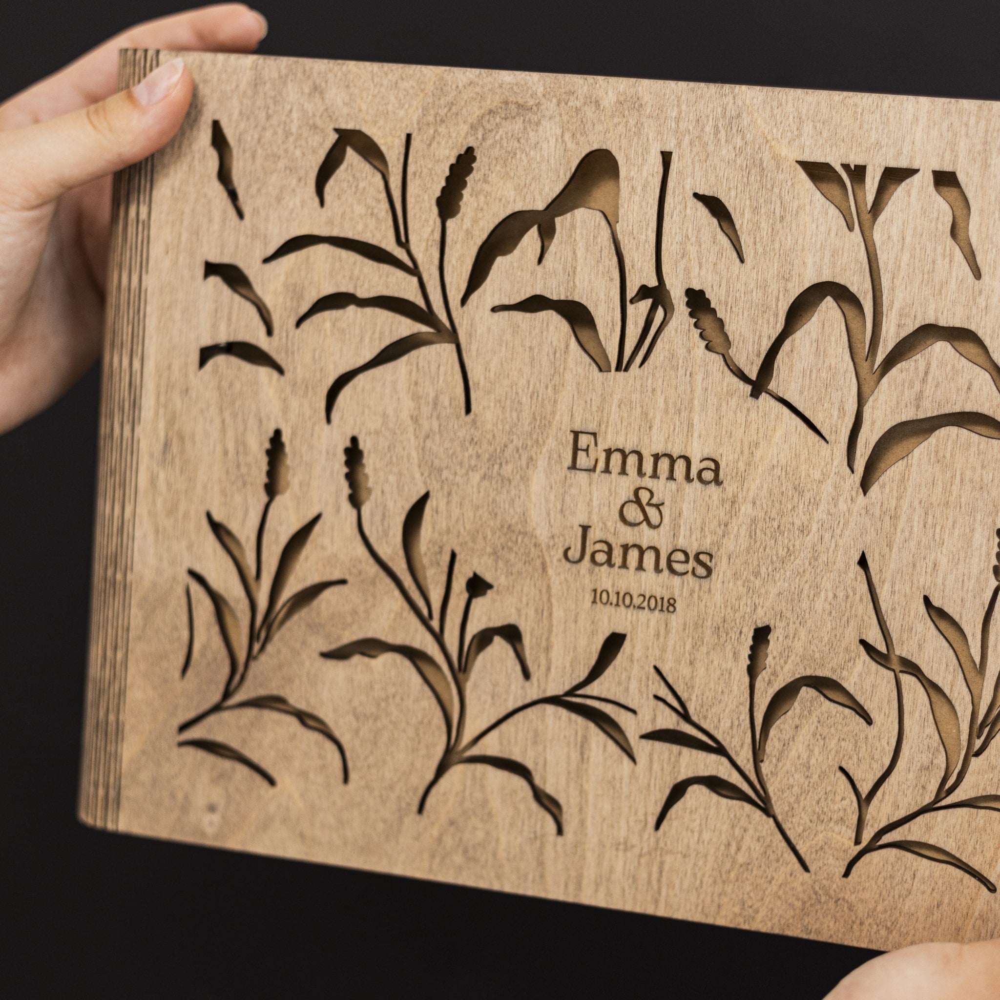 Wooden card with engraved floral designs and 'Emma & James' text on a dark background