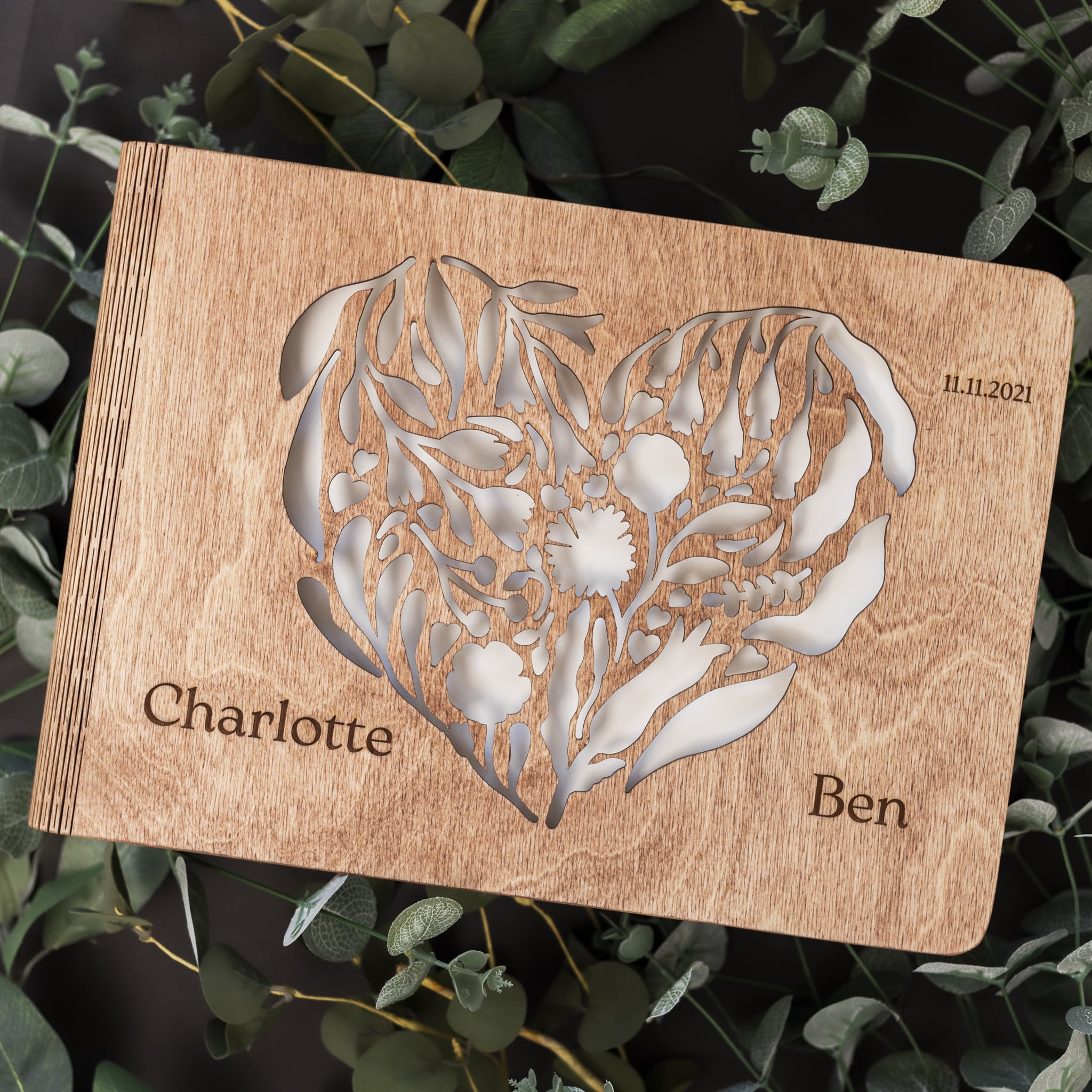 Wooden card with floral heart design and engraved names 'Charlotte' and 'Ben' on a leafy background.