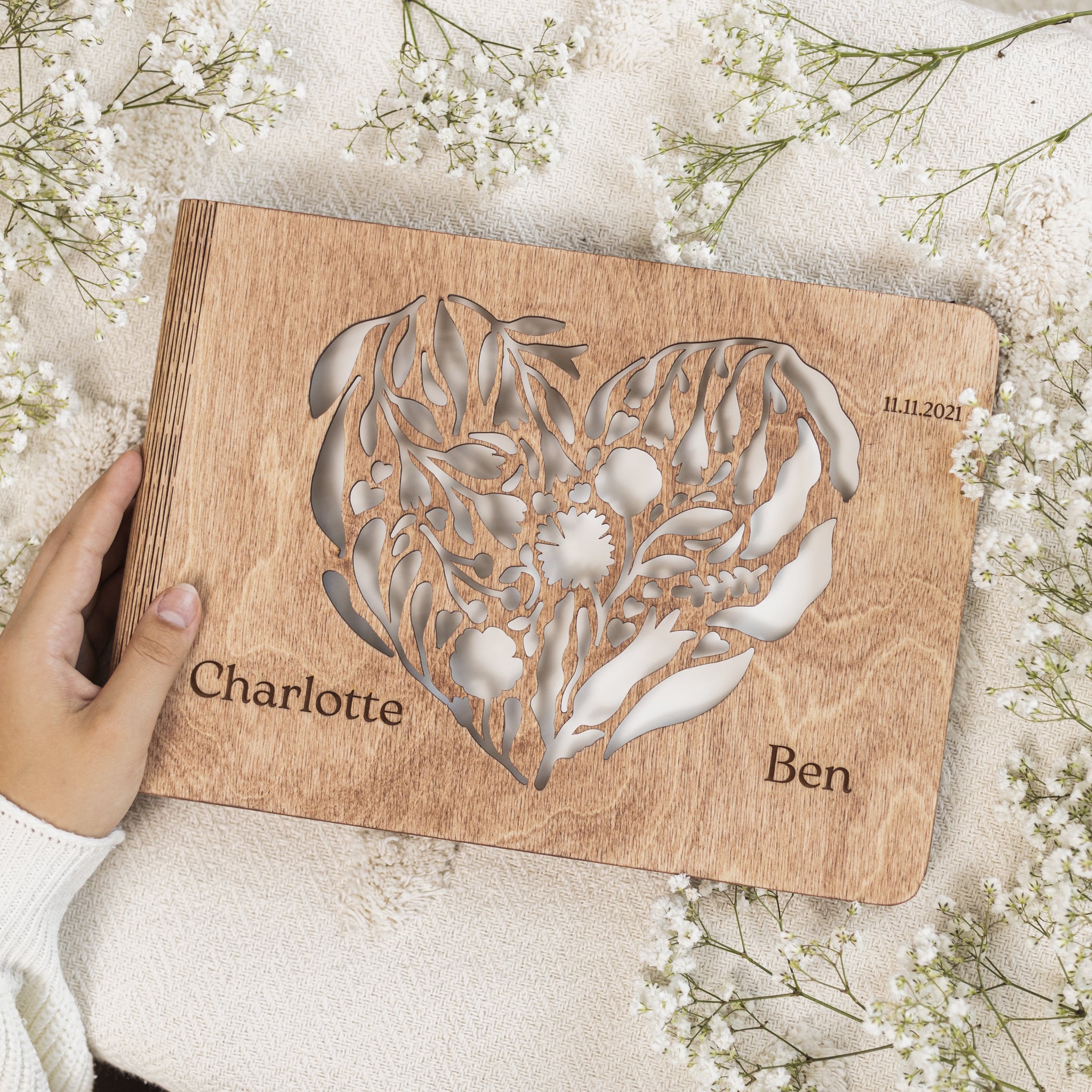 Wooden heart-shaped cutting board with engraved names and date, held by a hand on a floral background.