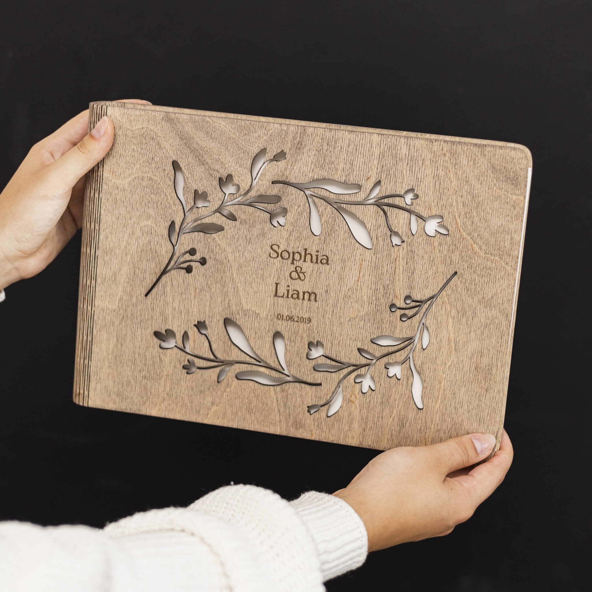 Wooden album with engraved floral design and text held by a person against a black background