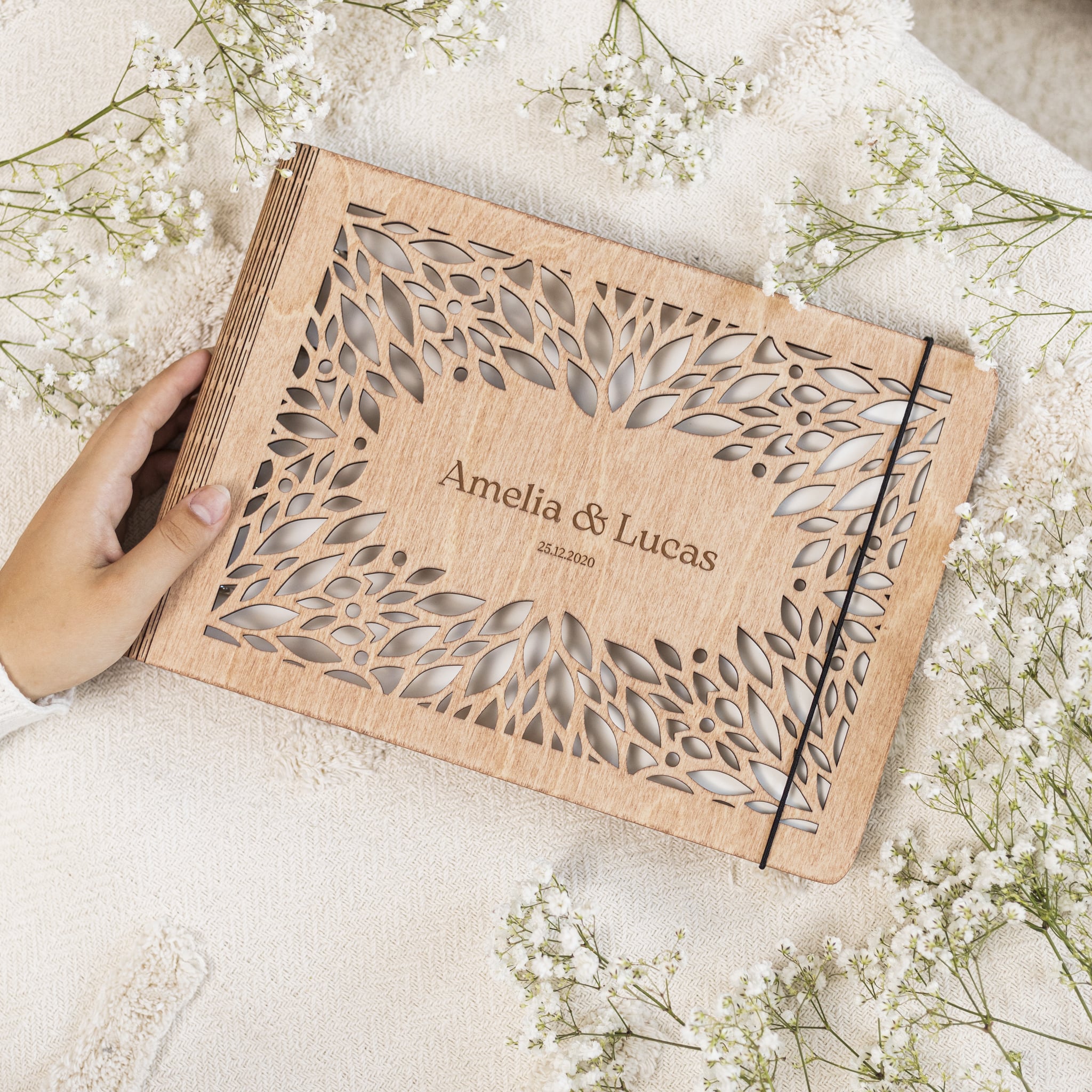 Wooden album with laser cut design and engraved names, held by a hand with white flowers in the background.