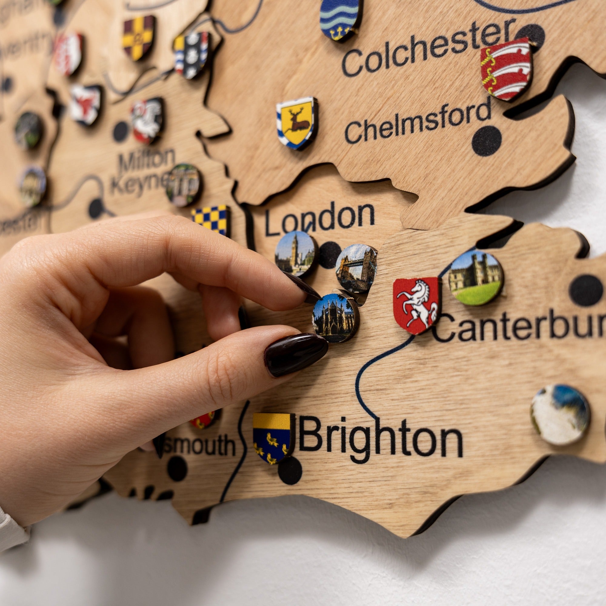 Wooden map of England with city names and symbols, hand placing a badge on 'Brighton'.