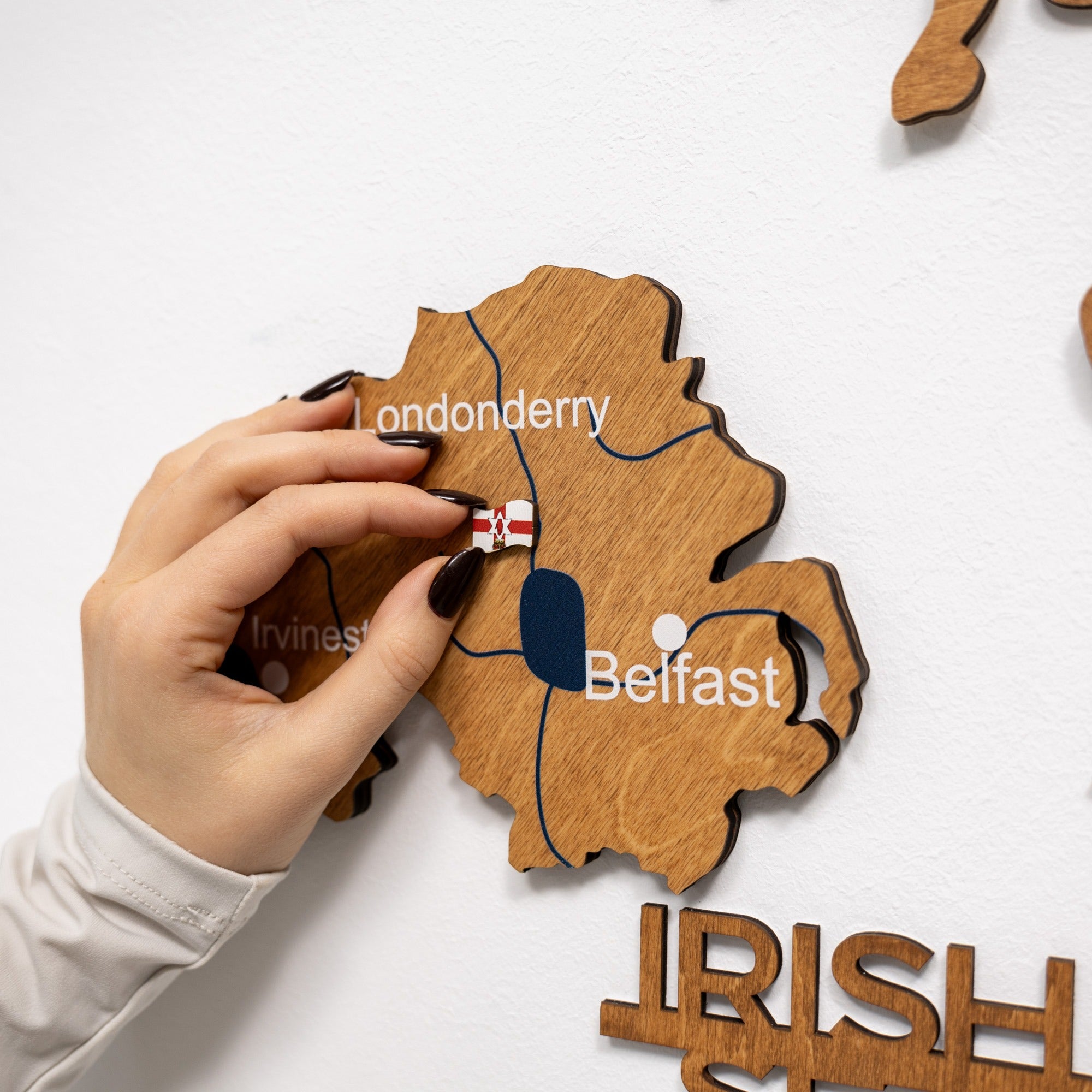 Wooden map of Ireland with Belfast labeled, hand holding a pin with Northern Ireland flag.