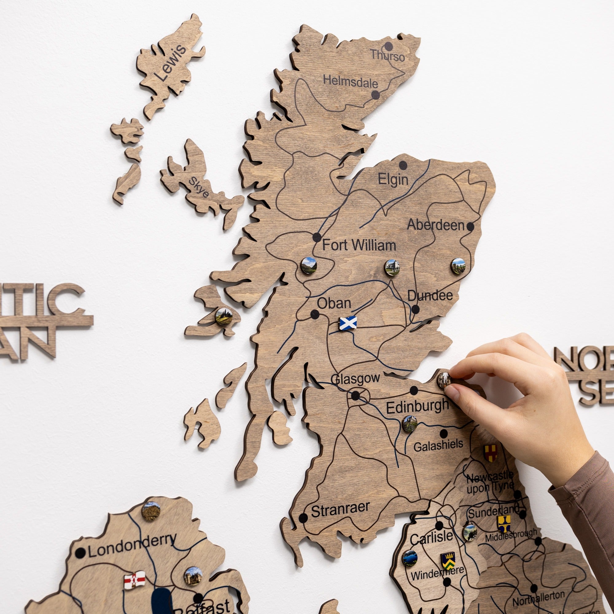 Wooden map of Scotland with cities labeled, held by a hand against a white background.