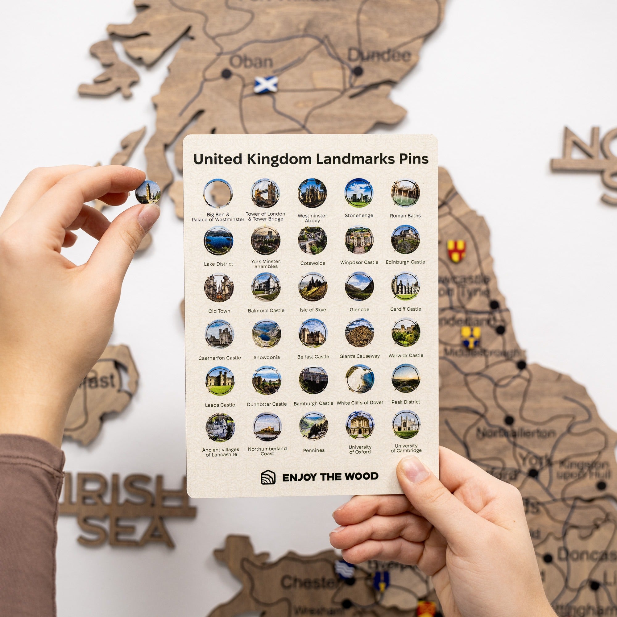 Sheet of United Kingdom landmarks pins held by a hand with a wooden map in the background.