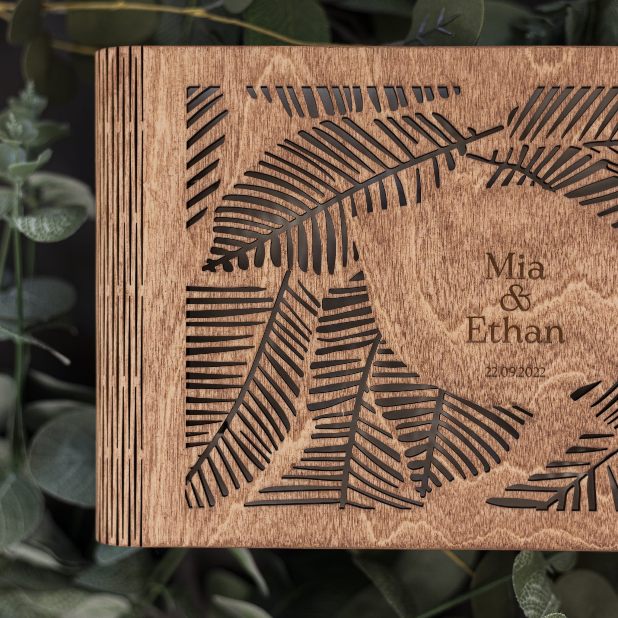 Wooden box with engraved leaf design and 'Mia & Ethan' text, surrounded by greenery.
