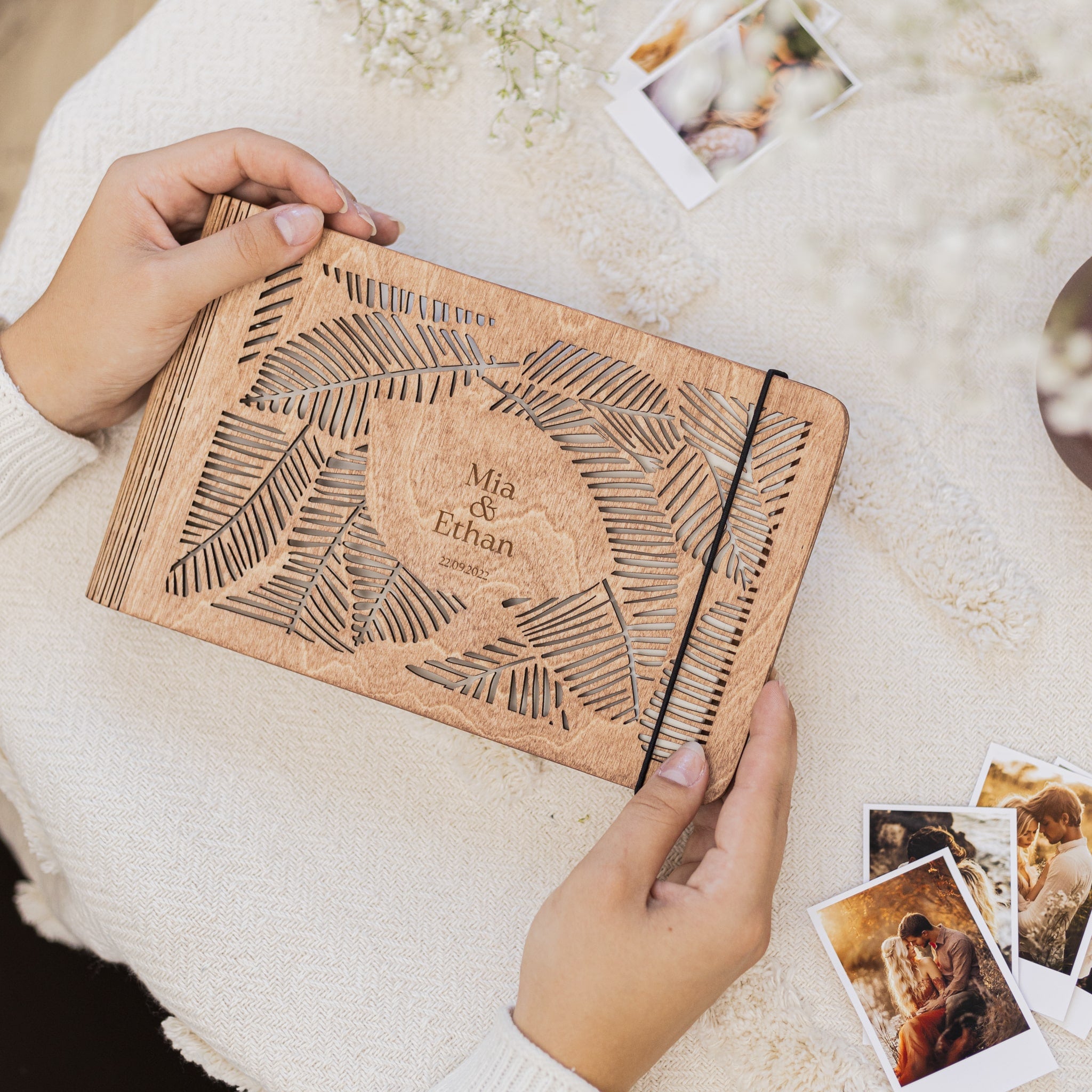 Person holding a wooden notebook with engraved design and text, surrounded by small photos on a light surface.