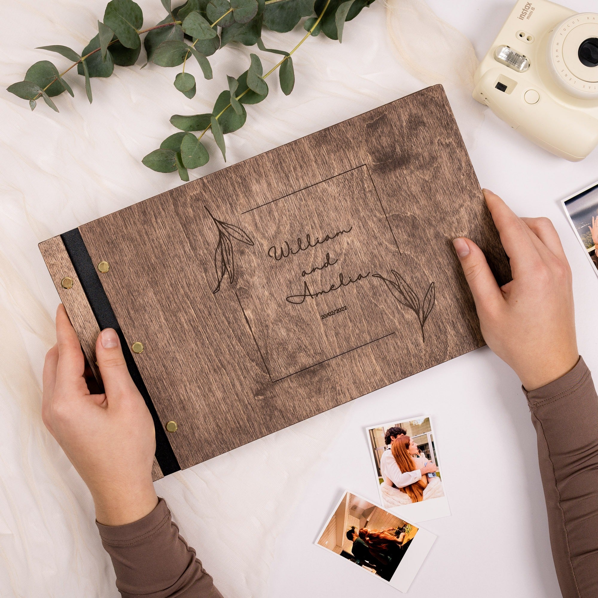 Wooden photo album with engraved text, held by hands on a white surface with greenery and a camera.