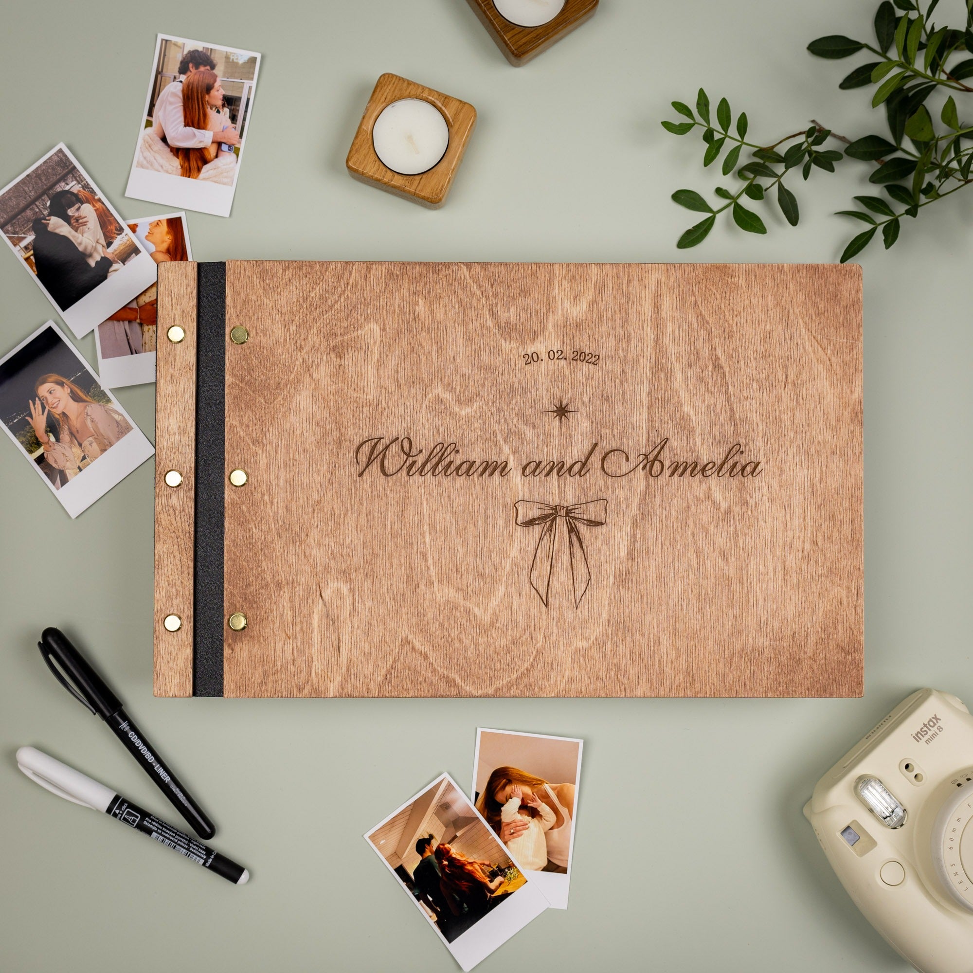 Wooden photo album with 'William and Amelia' engraving, surrounded by photos and a camera on a light green surface.