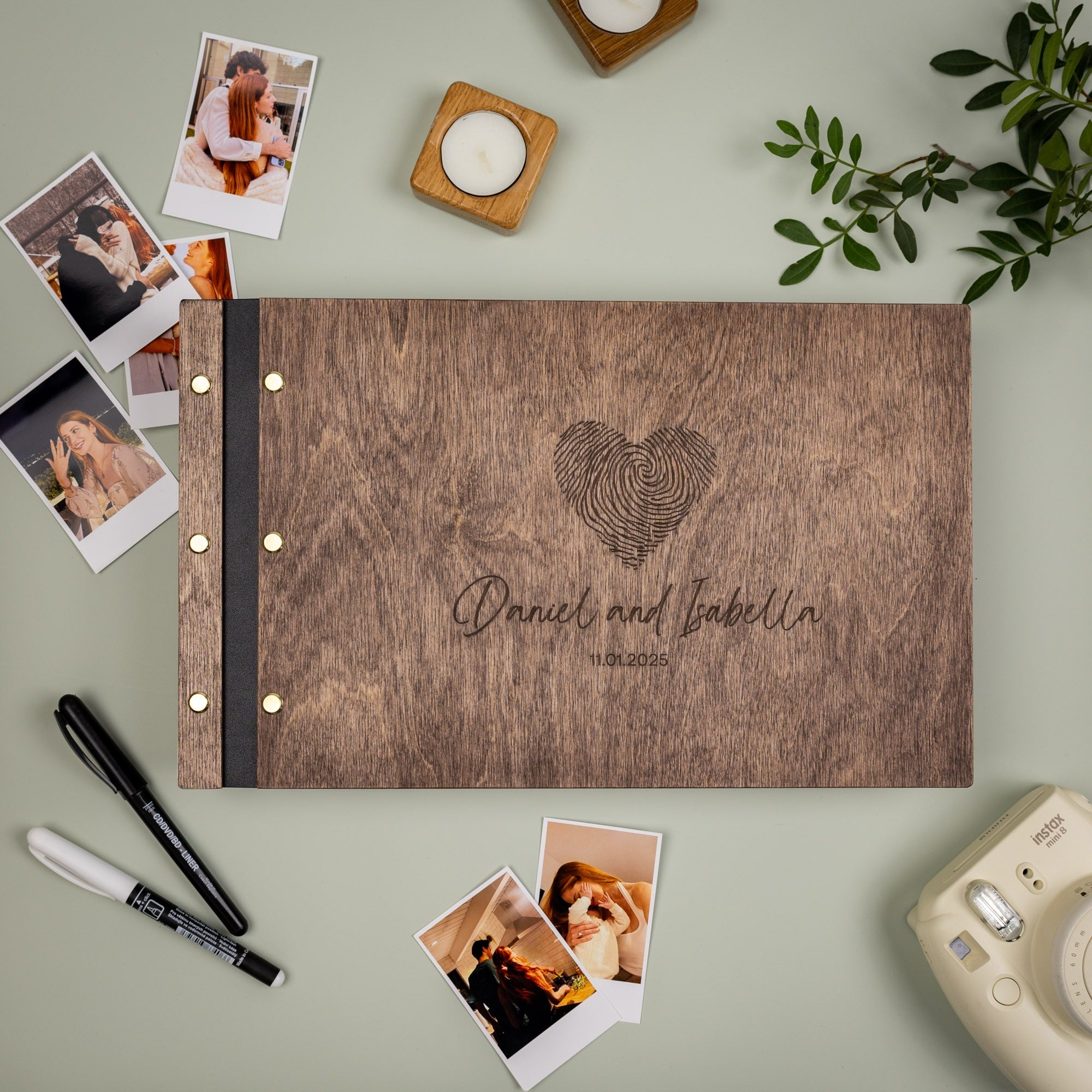 Wooden photo album with engraved heart design and text, surrounded by photos and a camera on a light green background.