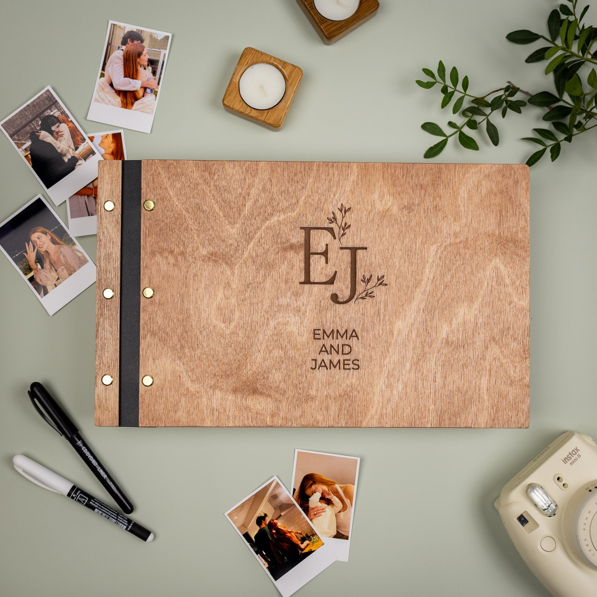 Wooden photo album with 'EJ' initials and 'Emma and James' text, surrounded by photos and a camera on a light green background.