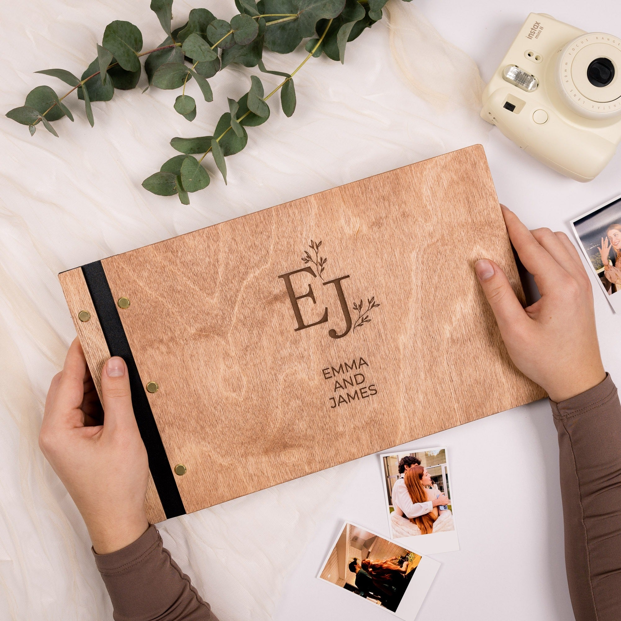 Personalized photo album with 'EJ' and 'Emma and James' on a white surface with greenery and a camera.