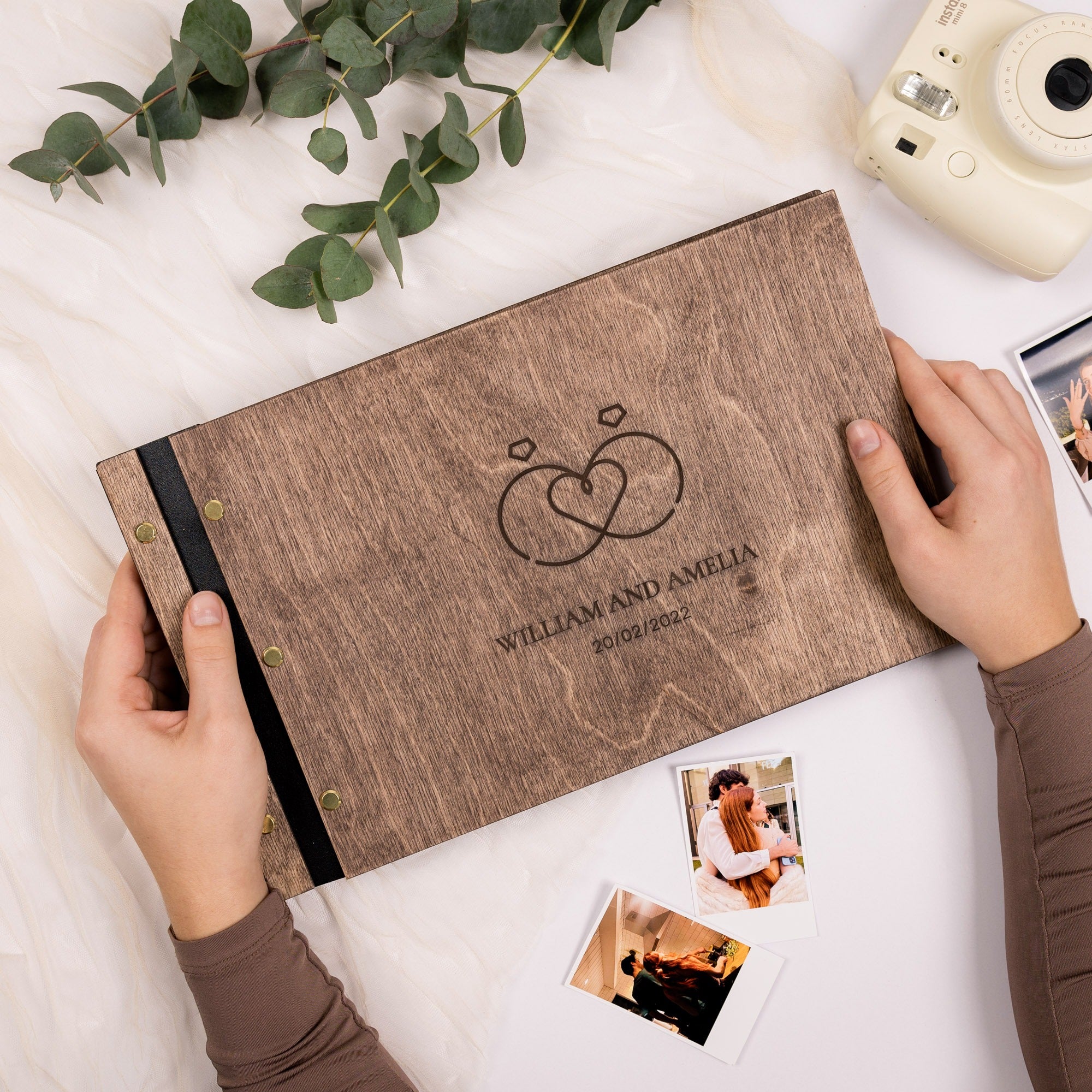 Personalized wooden photo album with hands holding it, surrounded by photos and greenery on a light surface.