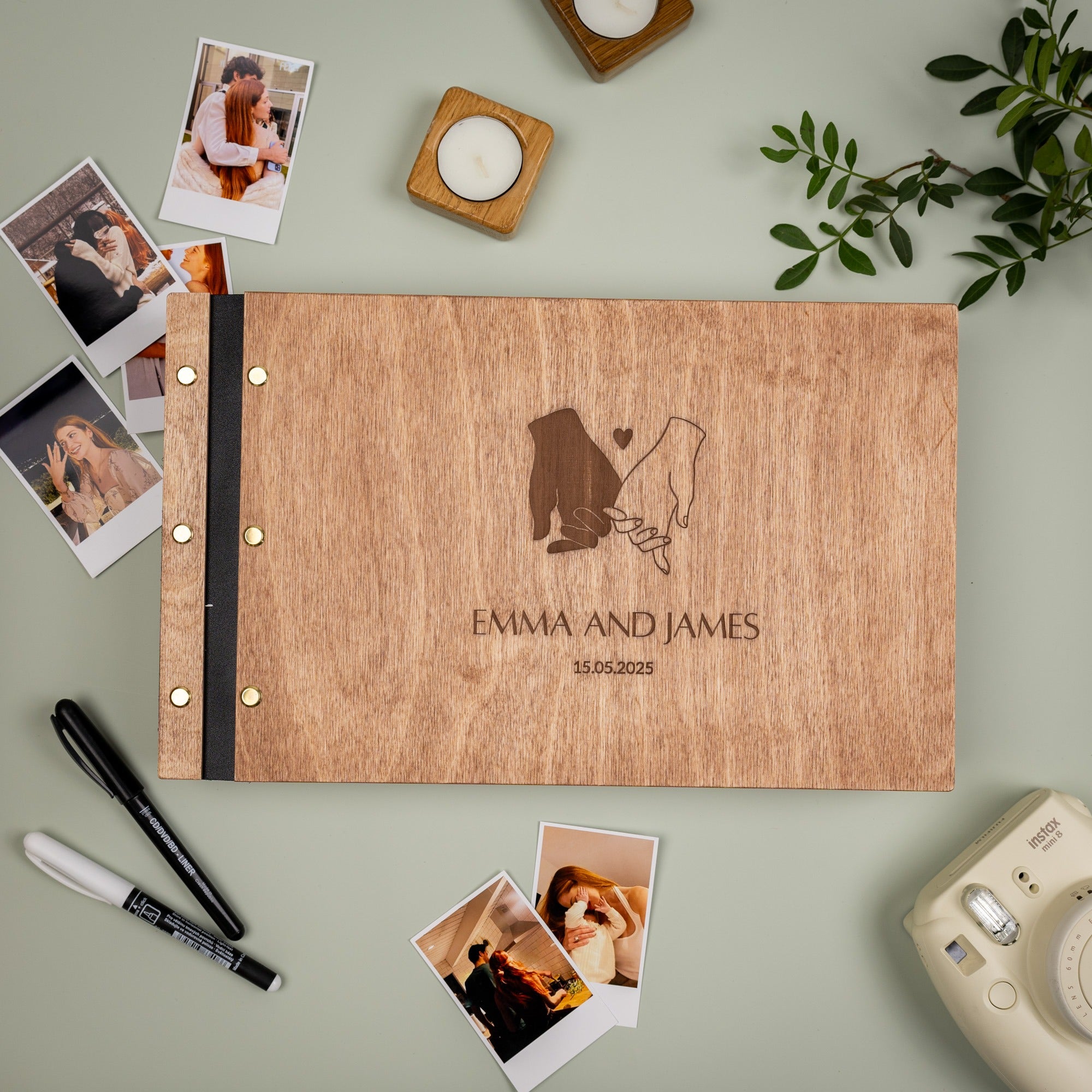 Wooden photo album with engraved couple and 'Emma and James' text, surrounded by photos and a camera on a light green background.