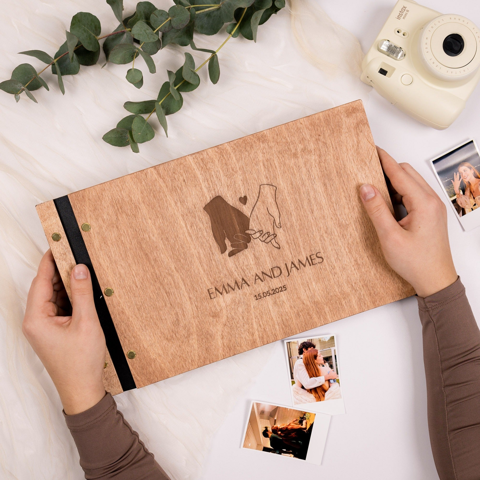 Wooden photo album with 'Emma and James' engraving, surrounded by photos and greenery on a white surface.