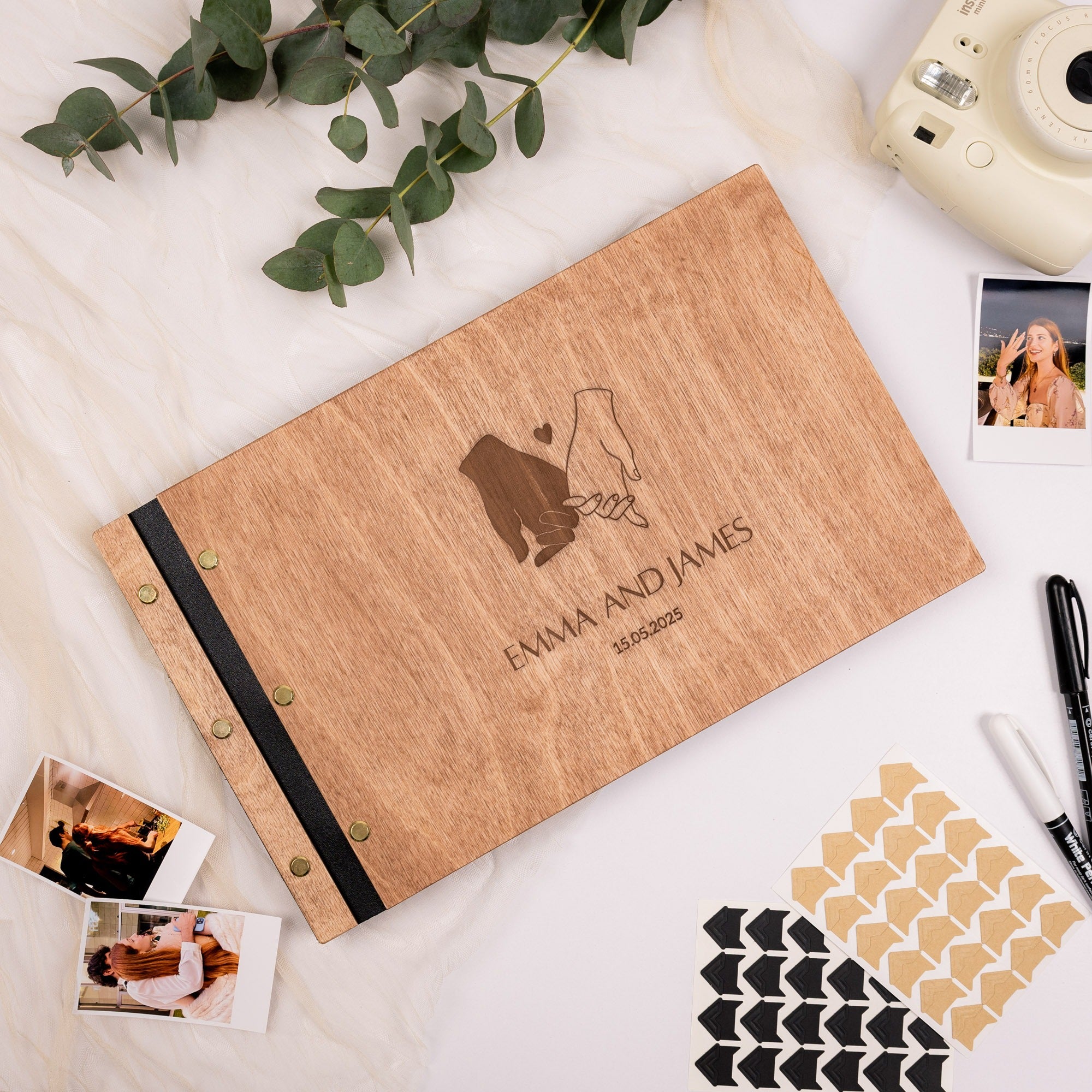 Wooden photo album with 'Emma and James' engraving on a light surface with photos and stationery items.