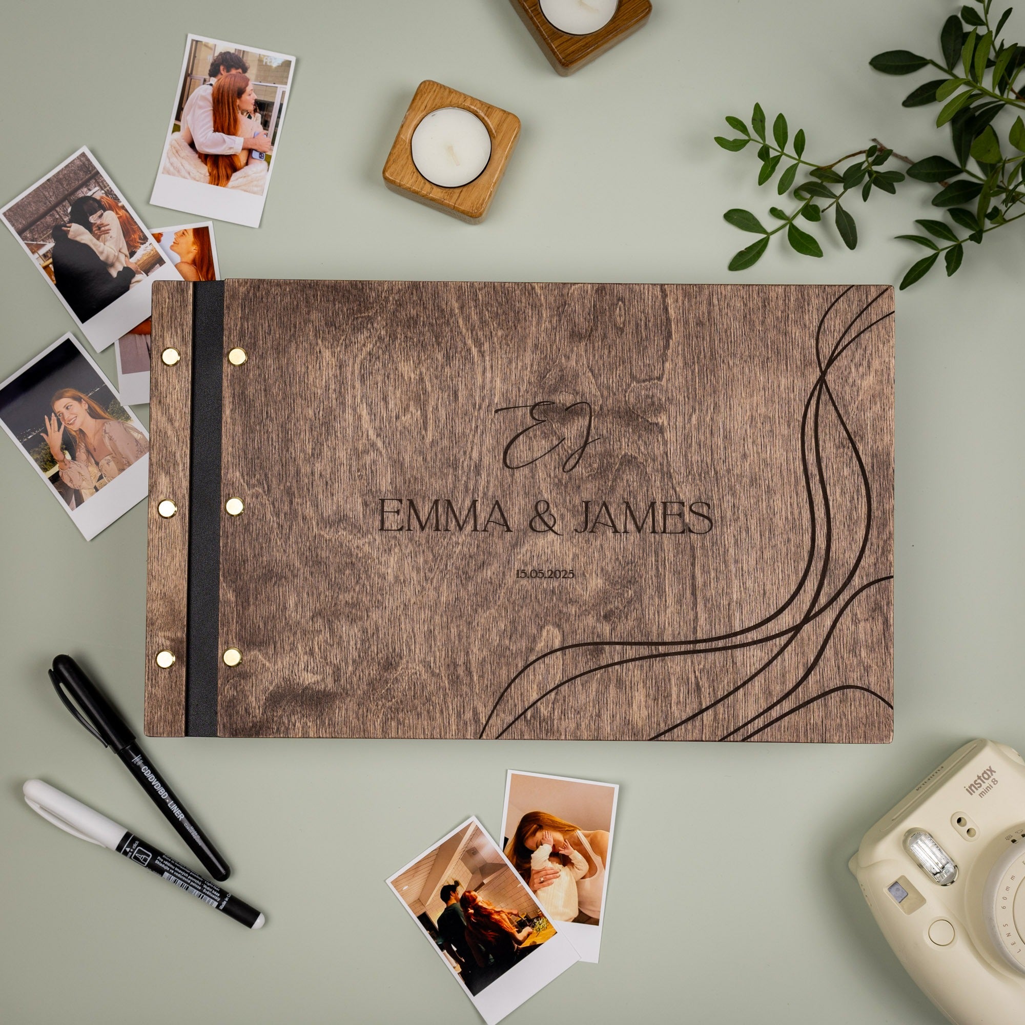 Wooden photo album with 'Emma & James' engraving, surrounded by photos and stationery on a light green surface.