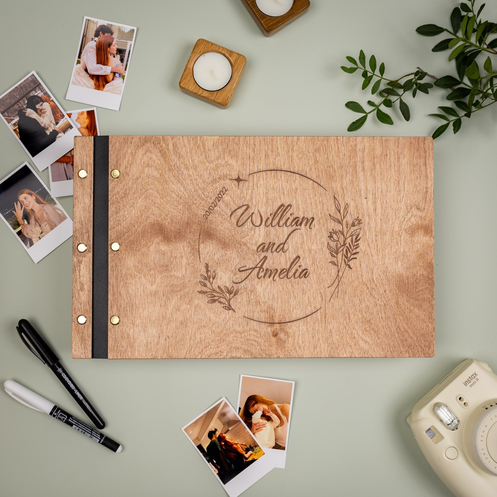 Wooden photo album with 'William and Amelia' engraving, surrounded by photos and a camera on a light green background.