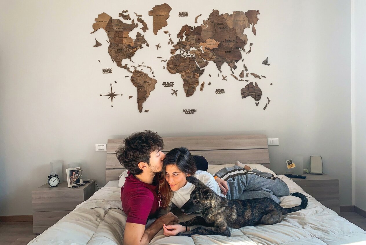 A couple cuddles with their dog on a bed beneath a world map