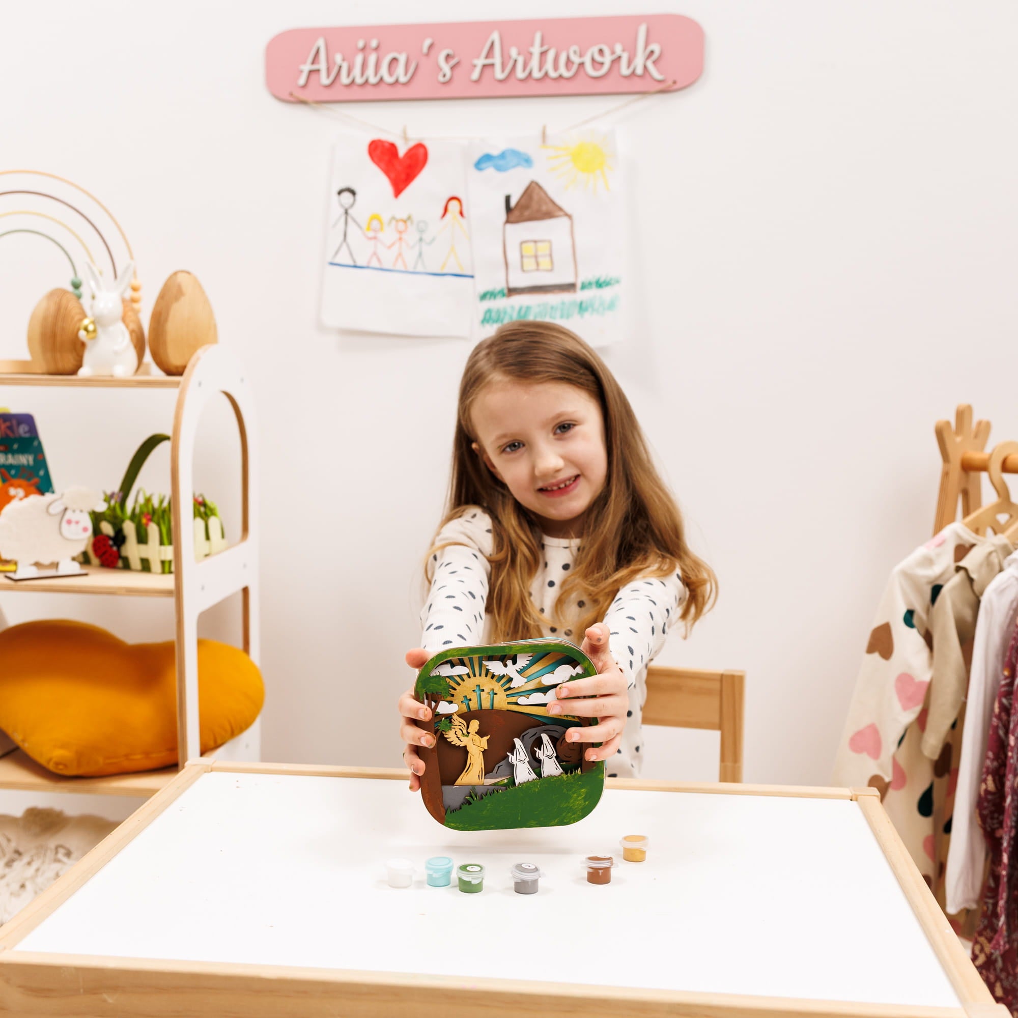 Young girl holding a small green box with artwork, sitting at a table in a room with shelves and children's items.