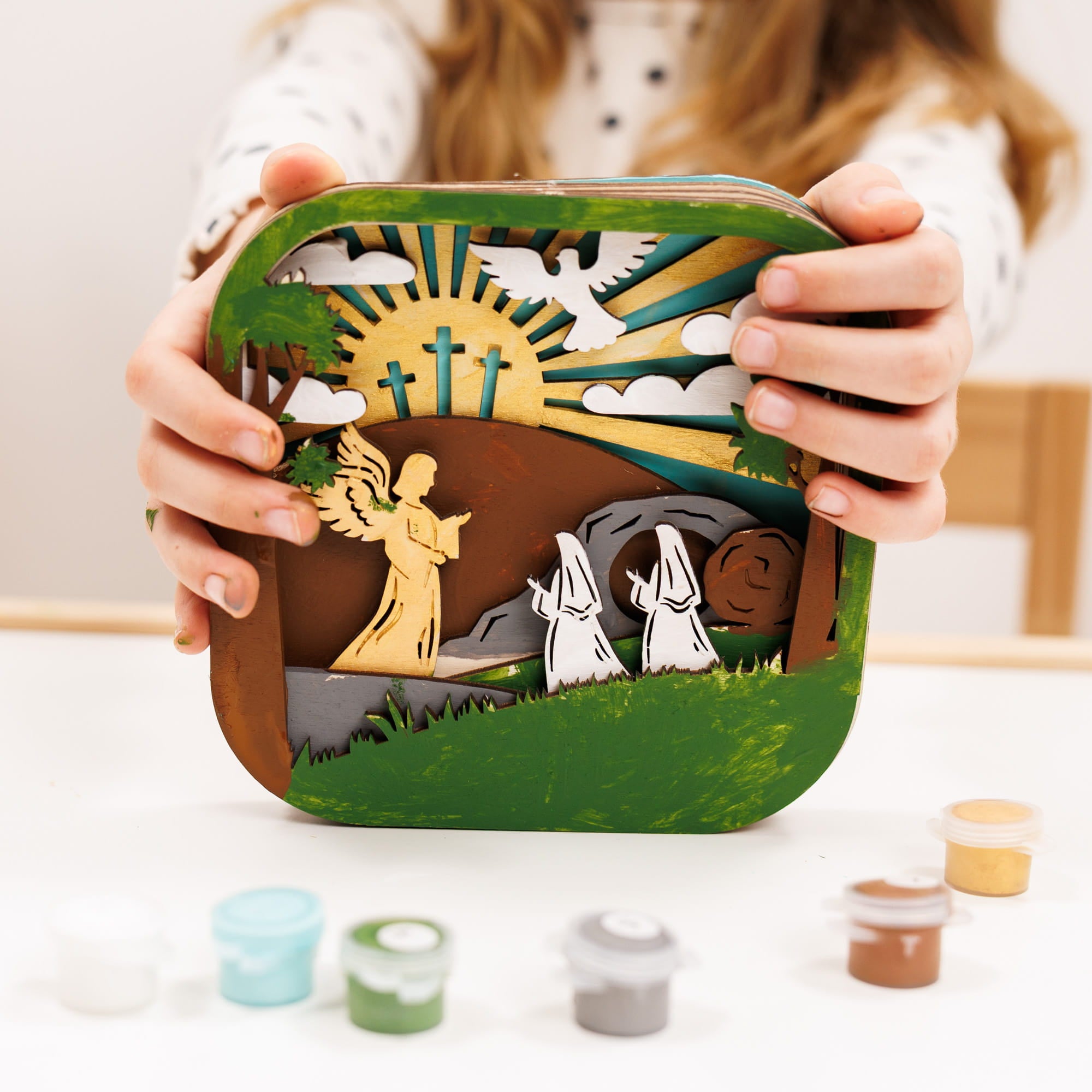 Hand holding a decorative wooden box with religious design, surrounded by paint containers.
