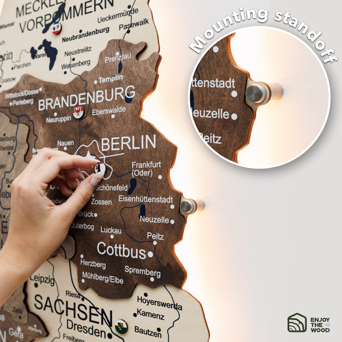 A wooden Germany wall map with illuminated interior lighting, shown mounted on a wall. A hand is pointing to Berlin, and a circular inset highlights the mounting hole.